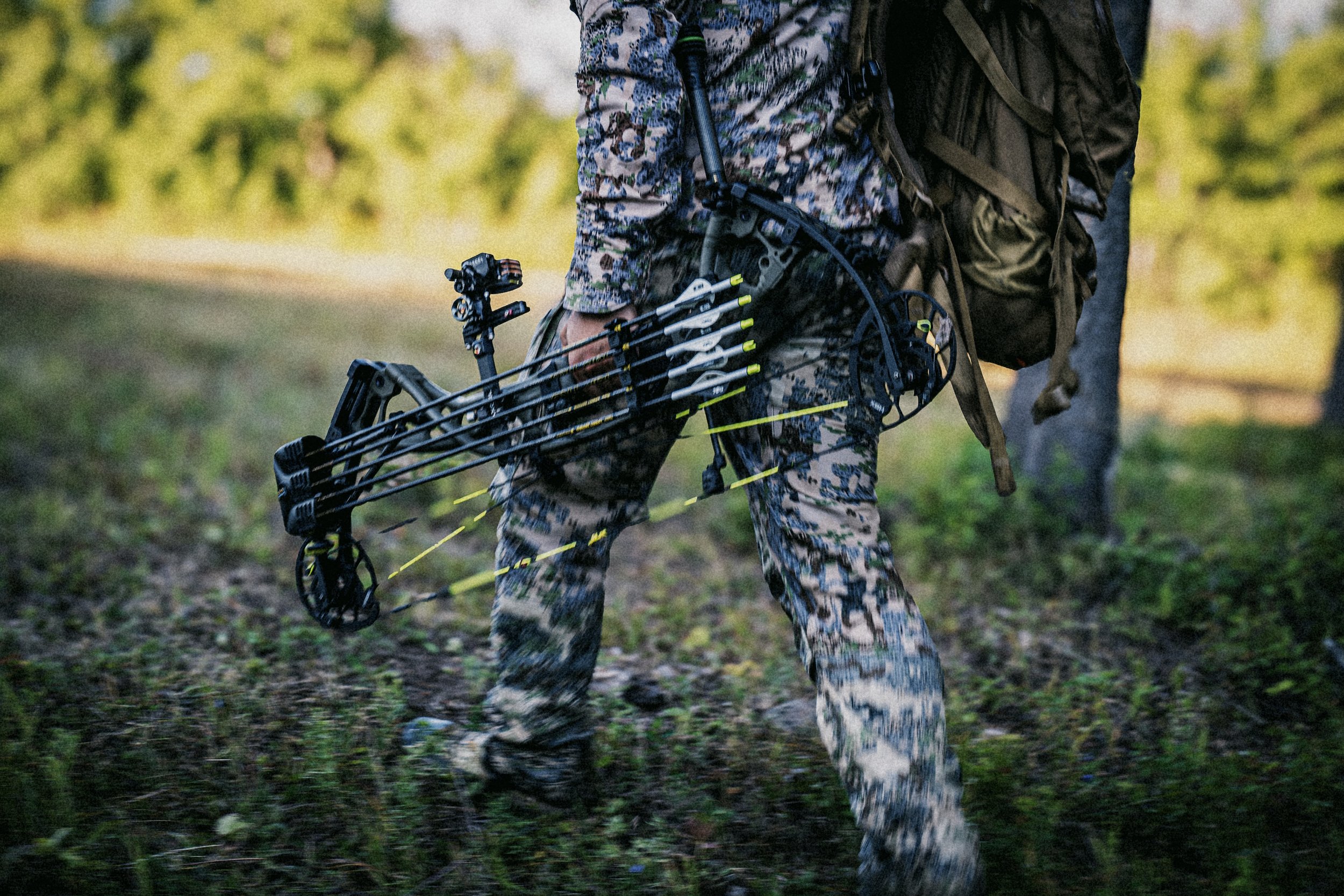 Person in camouflage clothing carrying a compound bow through a grassy wooded area. Product photoshoot.