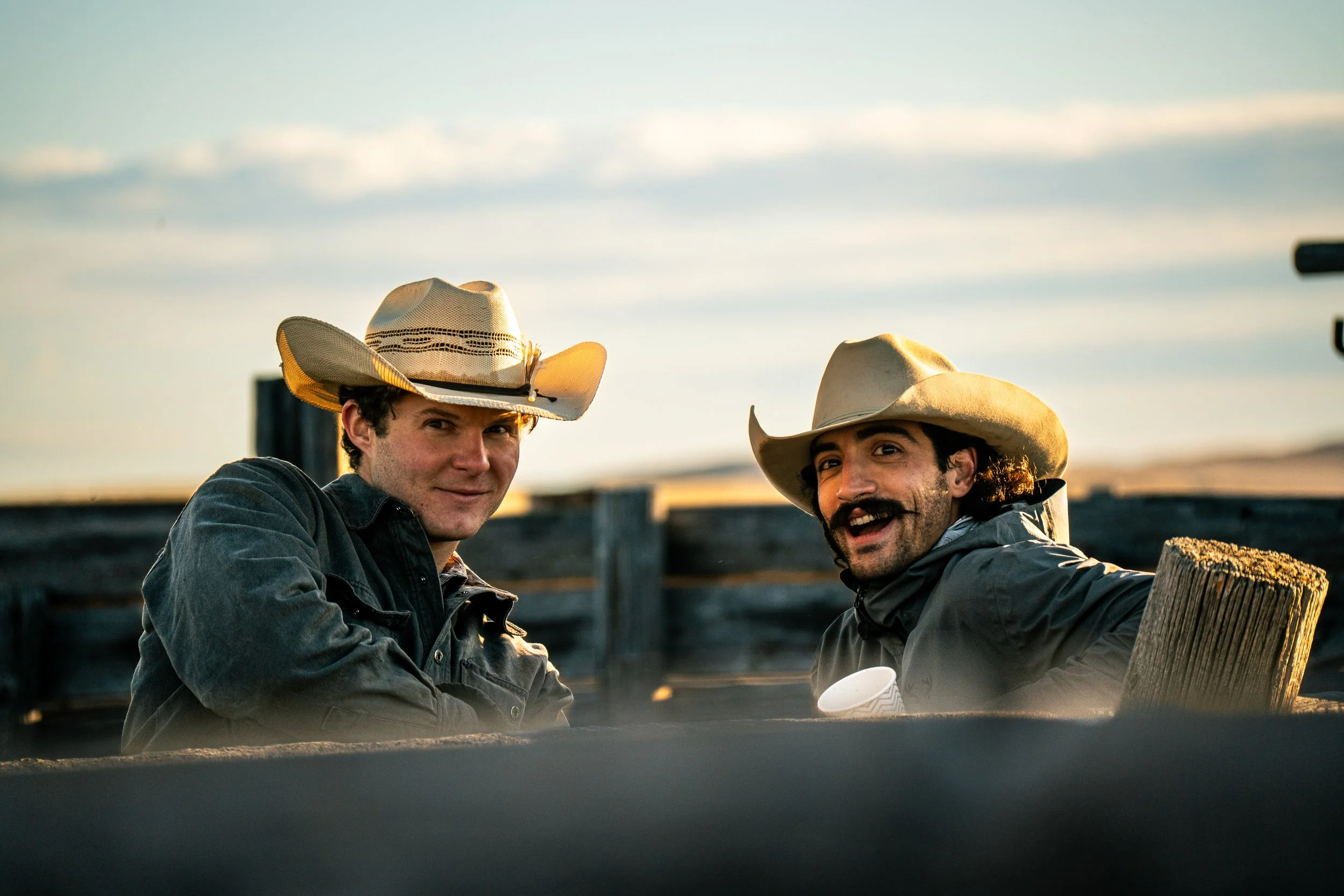 Two men wearing cowboy hats outdoors during sunset, smiling at the camera with a fence in the background.