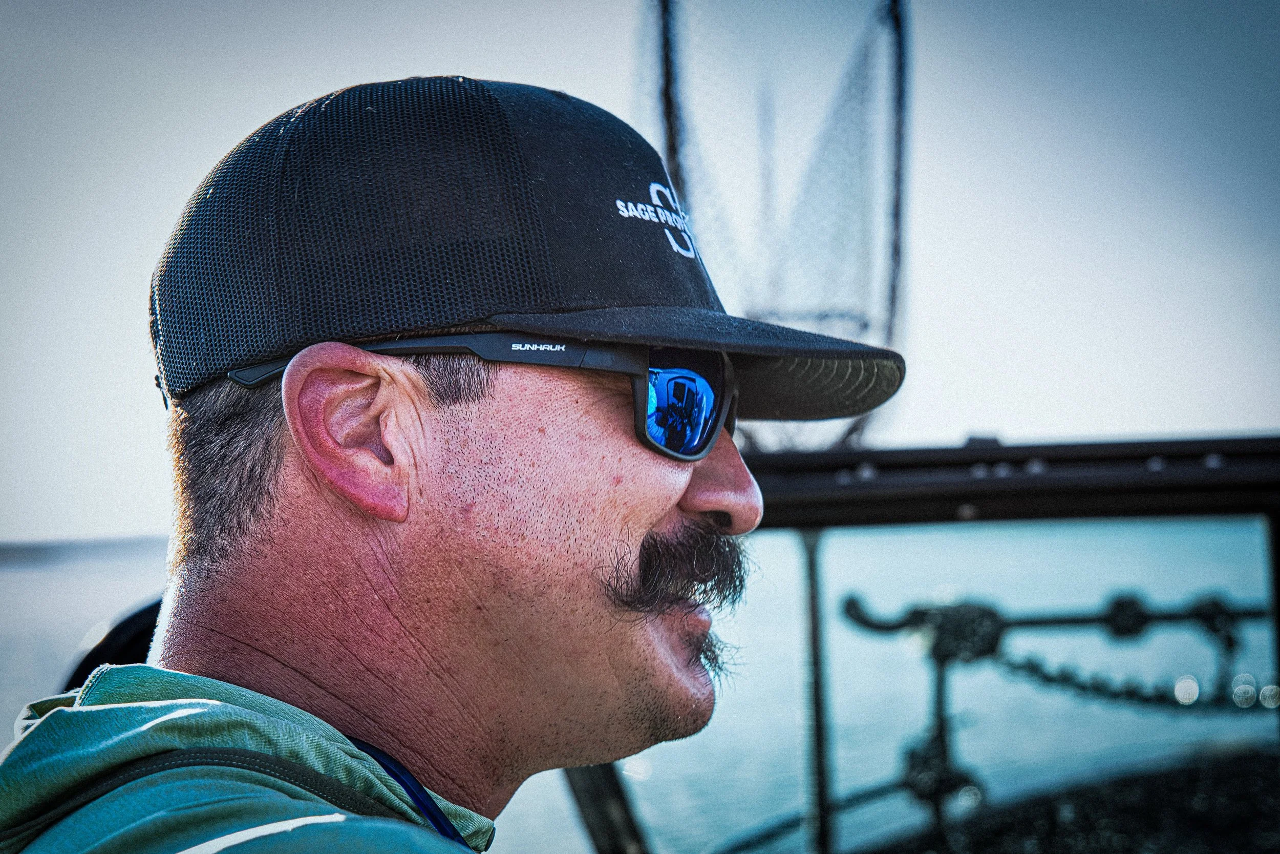 A man with a mustache and goatee wearing sunglasses, a black cap, and a hoodie, on a boat or near water. Fishing photoshoot.