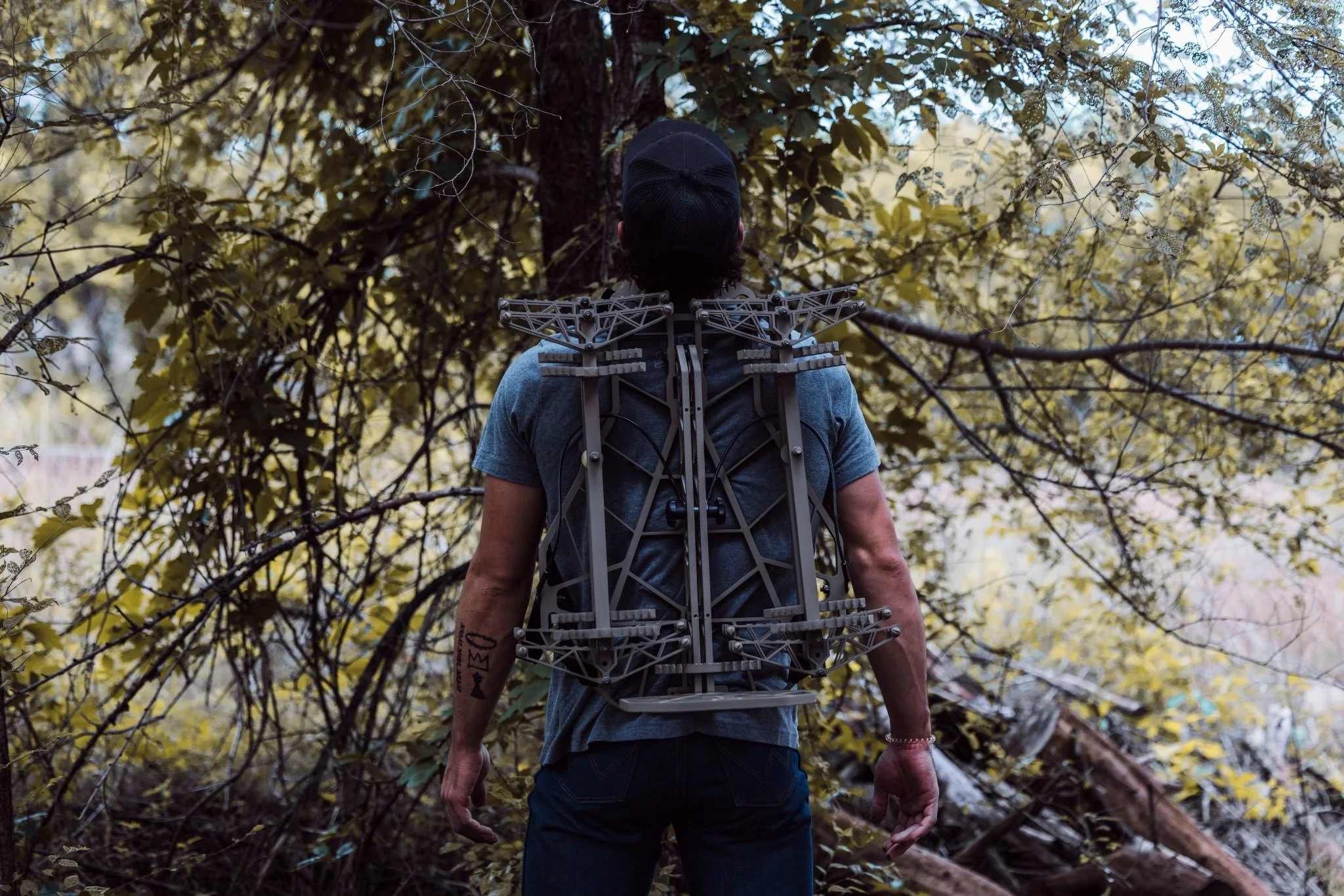 A person standing outdoors in a forest, back facing the camera, wearing a gray t-shirt, dark jeans, a black cap, and a large metallic exoskeleton on their back with multiple wires and mechanical parts amid trees and leaves. Tree Stand.
