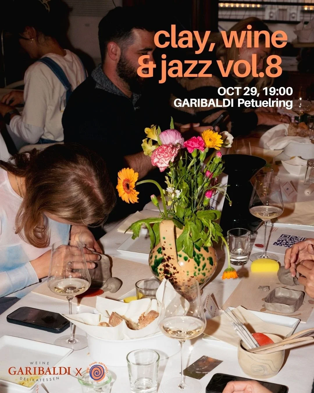 People gathered around a dining table with a colorful floral centerpiece, wine glasses, and plates, at a wine and jazz event.