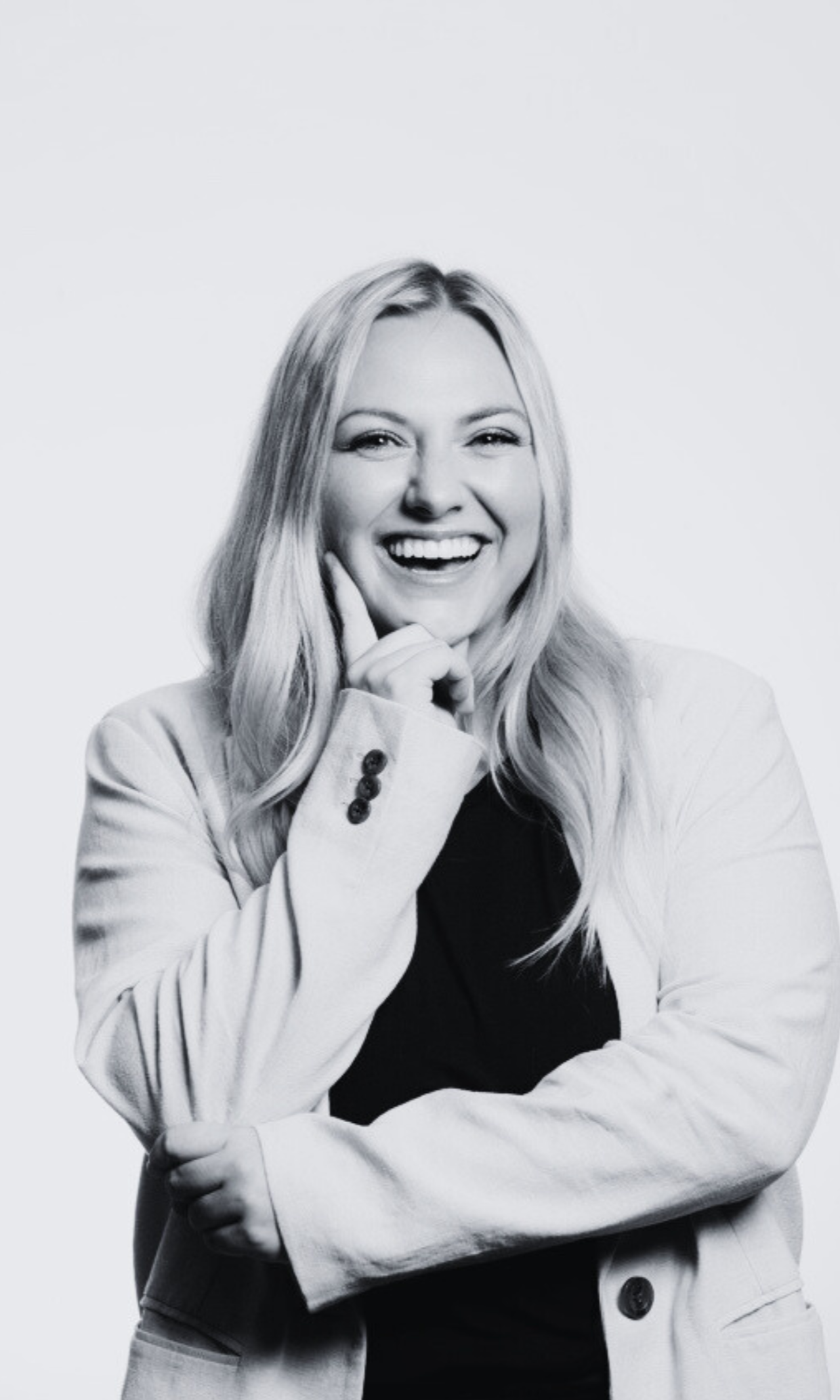 Black and white photo of a woman smiling, with her left hand touching her face and her right arm crossed, wearing a light-colored blazer over a dark top.