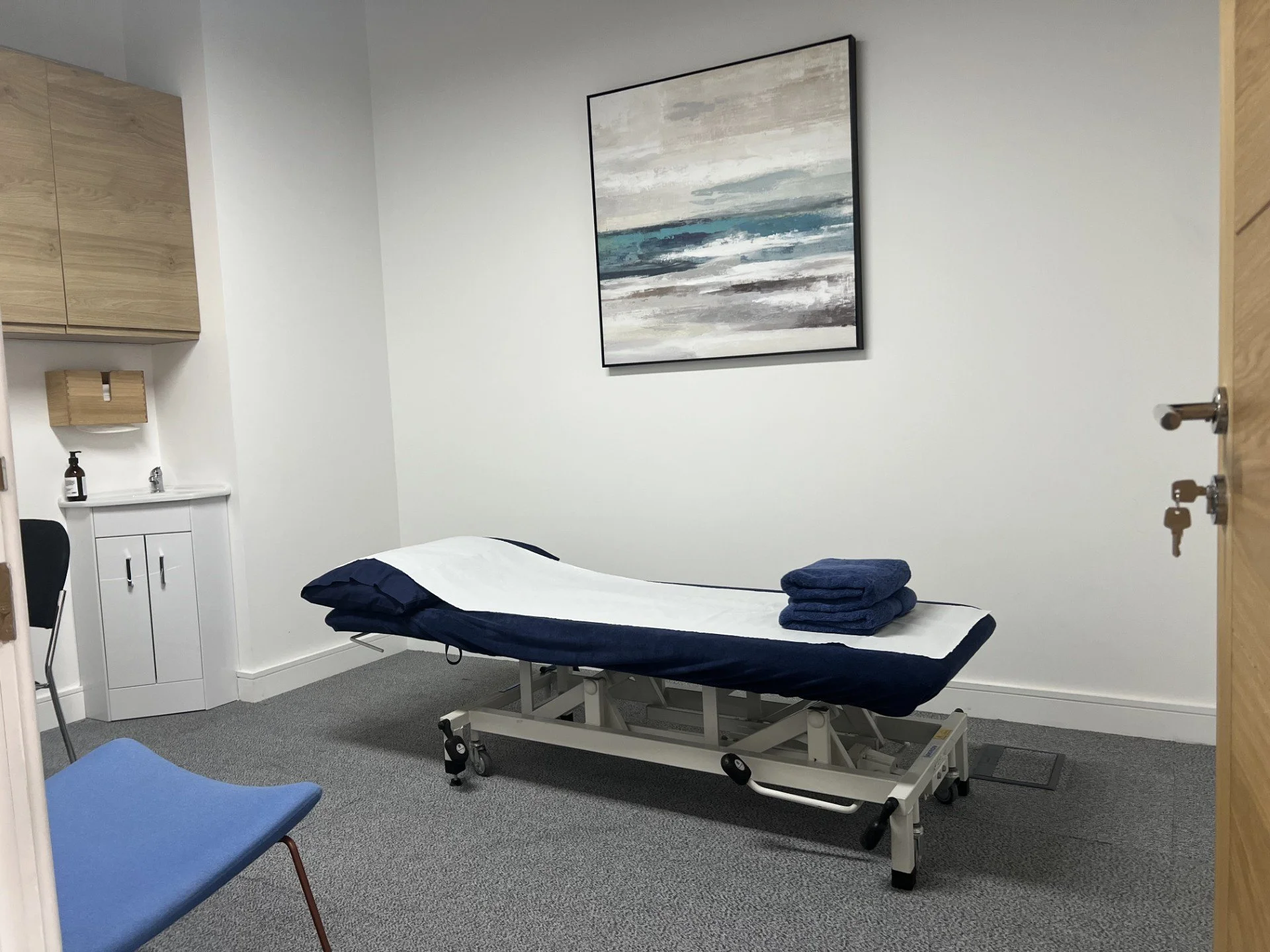 A medical examination room with a bed covered in a white sheet and two folded navy blue towels on top. There is a wooden cabinet with a soap dispenser next to a small sink. The room has gray carpeted flooring, a blue chair, and a large abstract painting on the white wall.
