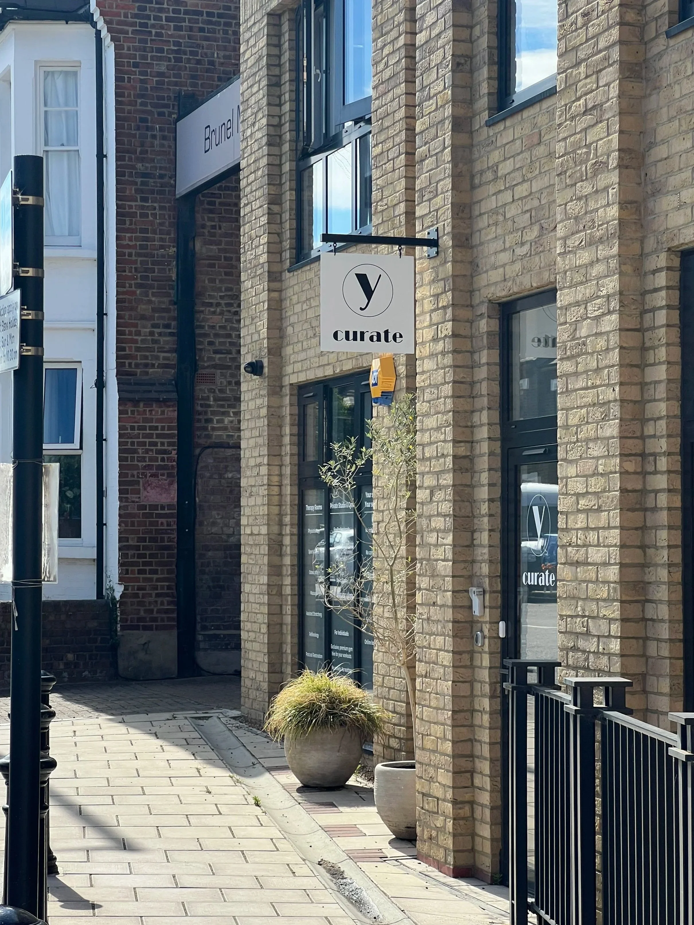Shop front view of private hire wellbeing hub, 'y curate', in Kilburn Lane, W10, hanging outside, some potted plants, and large windows reflecting the sky.