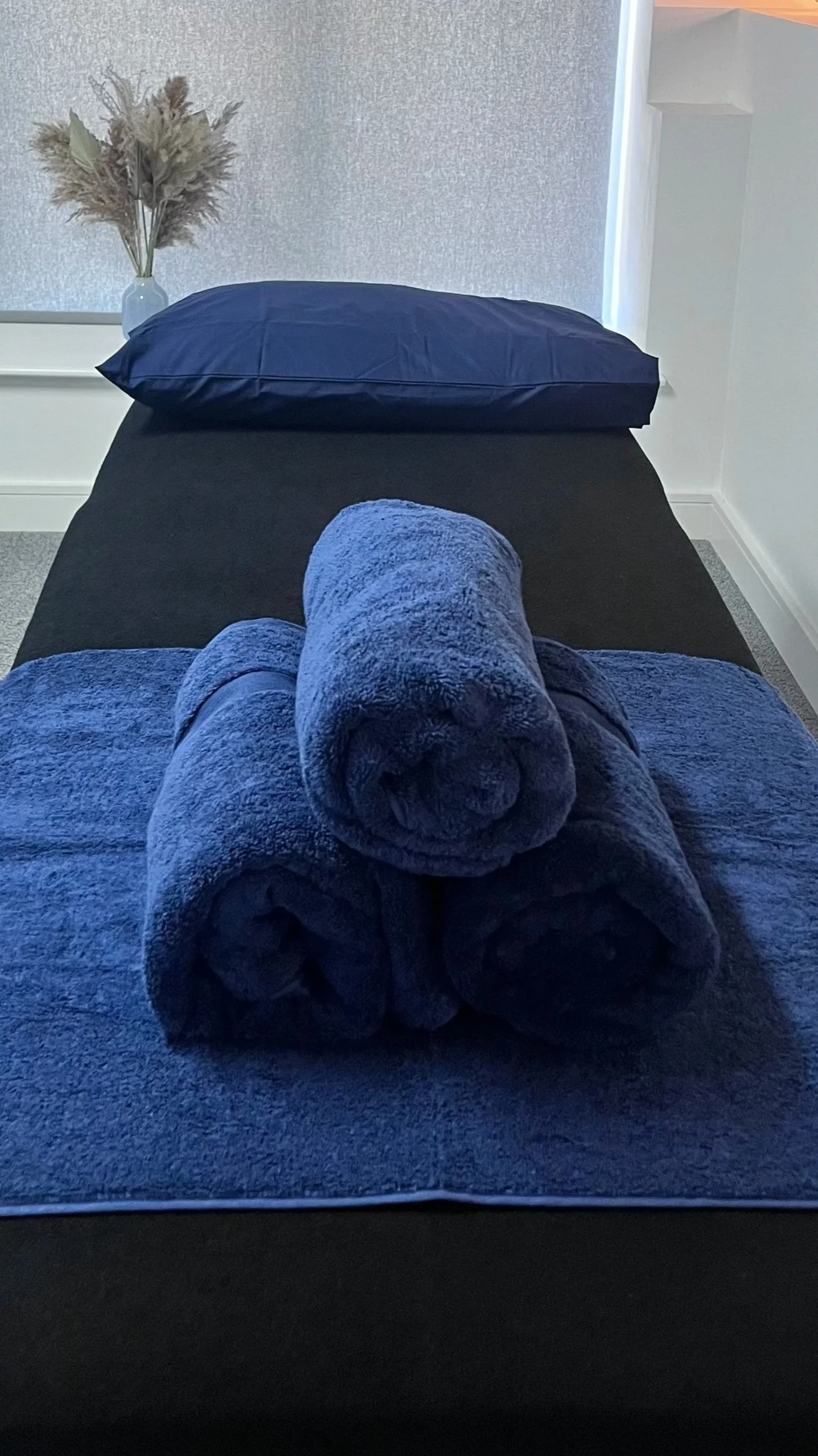 A massage or spa treatment room with a massage table draped in dark fabric, with three rolled towels on top. There is a pillow at the head of the table, and a vase with dried pampas grass on a small table or shelf nearby. The room has textured wallpaper, a window with roller shade, and soft lighting.