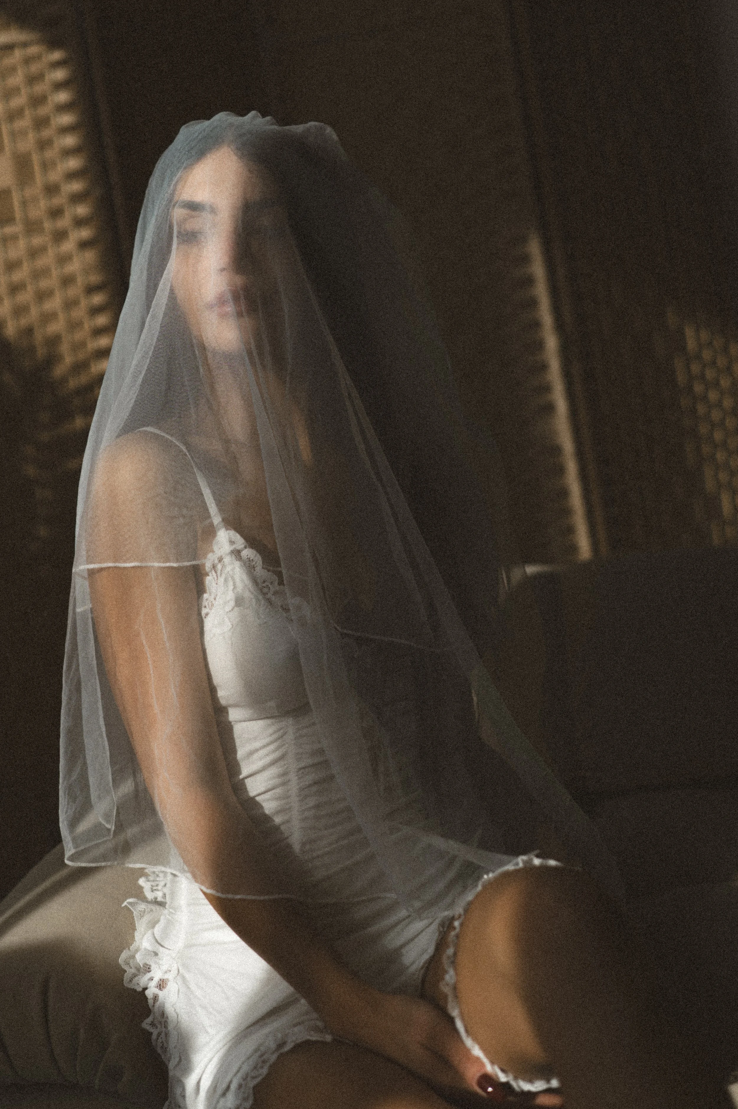 A woman with dark hair and light skin, wearing a white lace camisole and shorts, sitting on a beige chair with a sheer veil covering her face and head, against a dark wooden background.