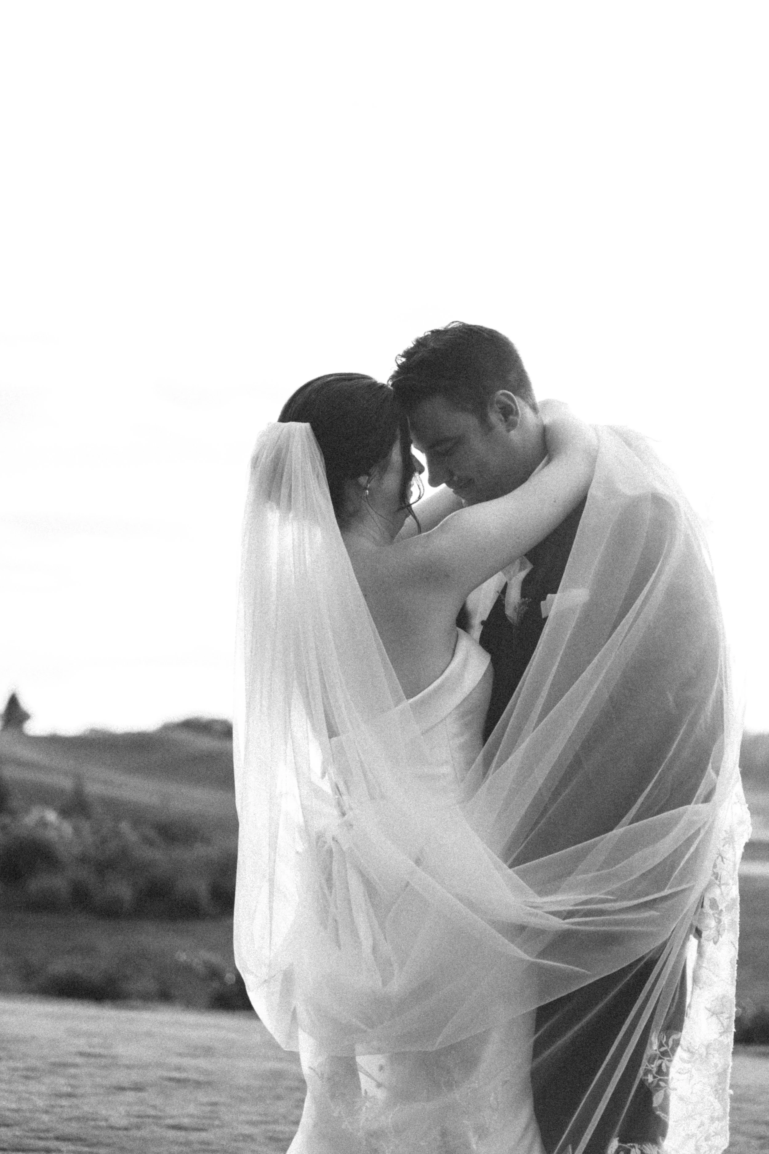 Black and white photo of a bride and groom embracing outdoors, the bride wearing a veil and dress, and the groom in a suit, with their foreheads touching.