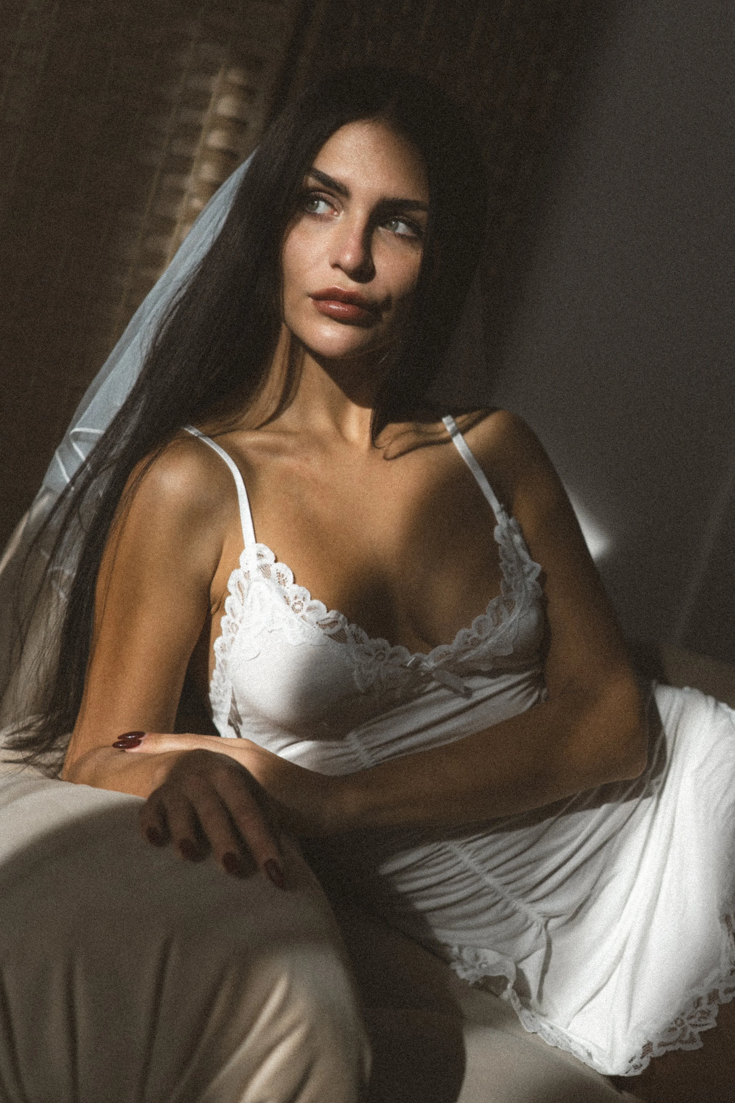 A woman with long dark hair and light skin, wearing a white lace-trimmed camisole, sitting on a couch with sunlight casting shadows across her face and body.
