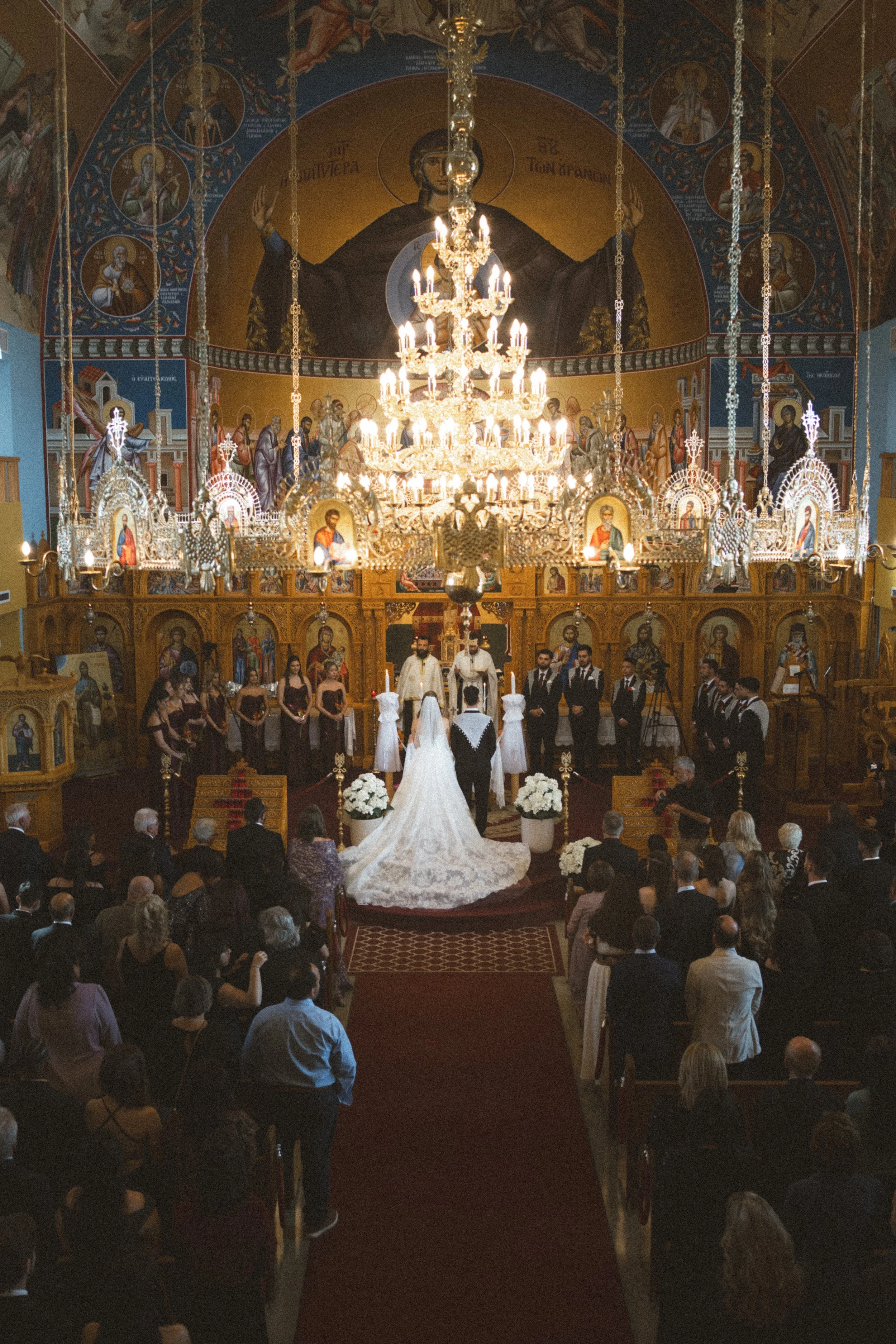 A wedding ceremony inside a church with a large chandelier, icons, and religious artwork. The bride and groom are at the altar with witnesses and guests present.