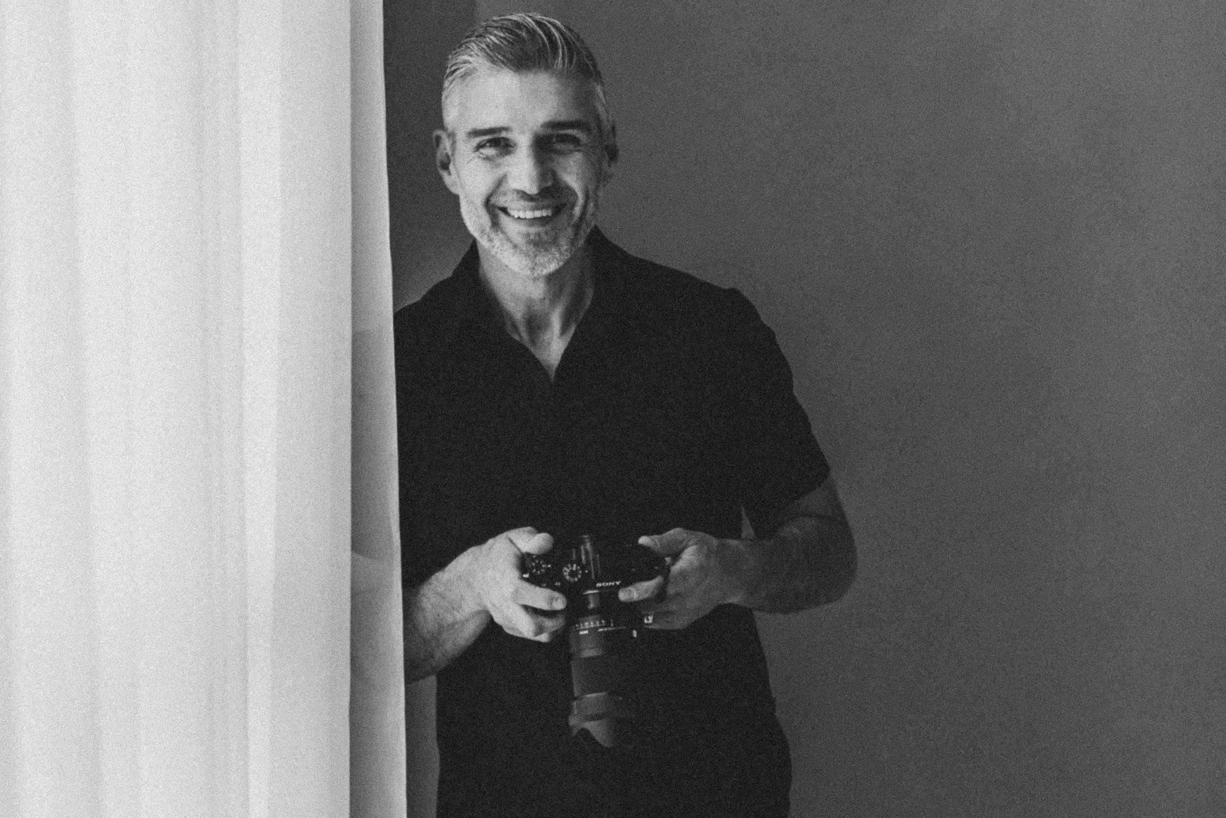 A smiling man with gray hair and a beard holding a camera, partially behind a curtain, in a black and white photo.