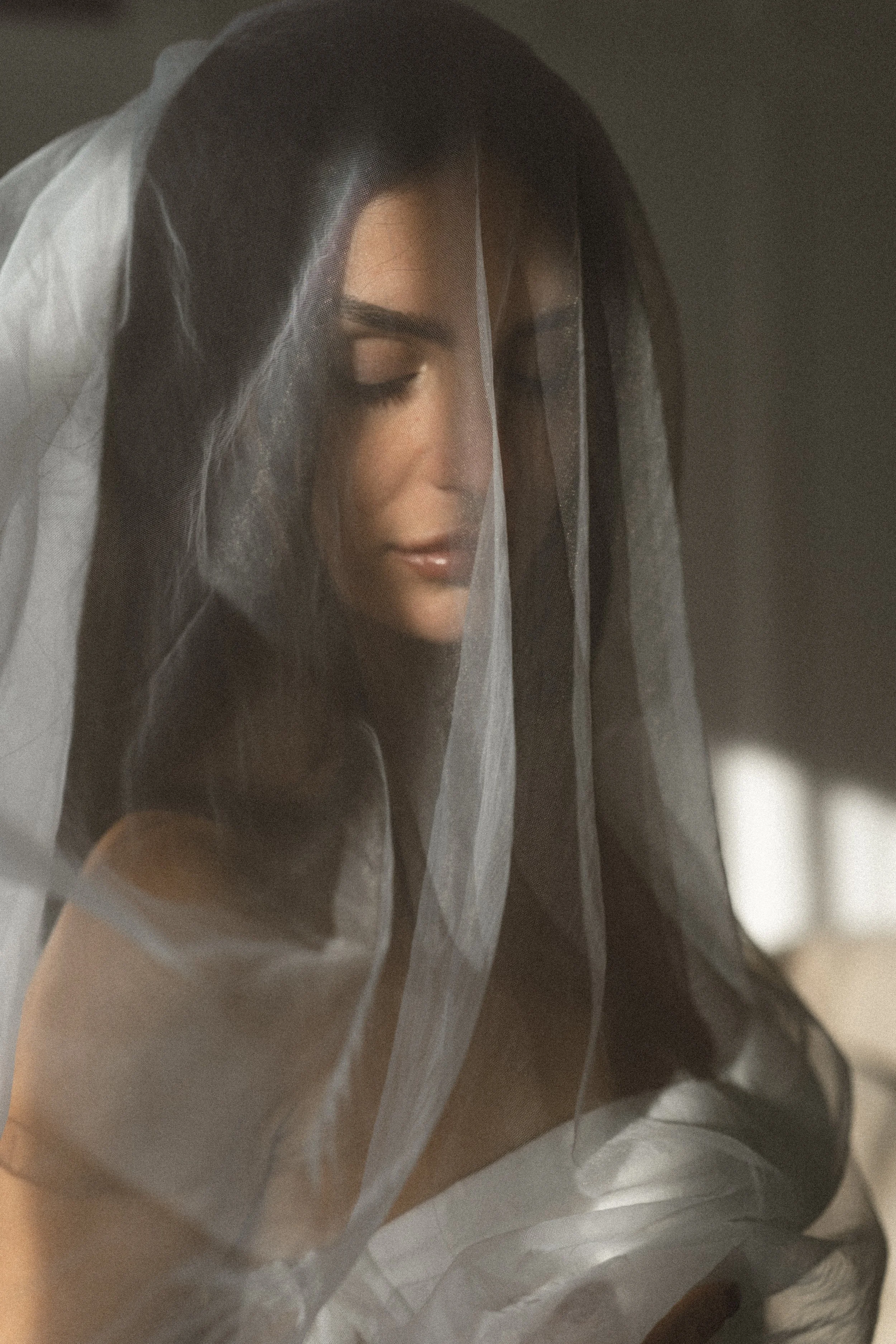 A woman with dark hair and closed eyes, partially covered with a sheer white veil, in a softly lit room.