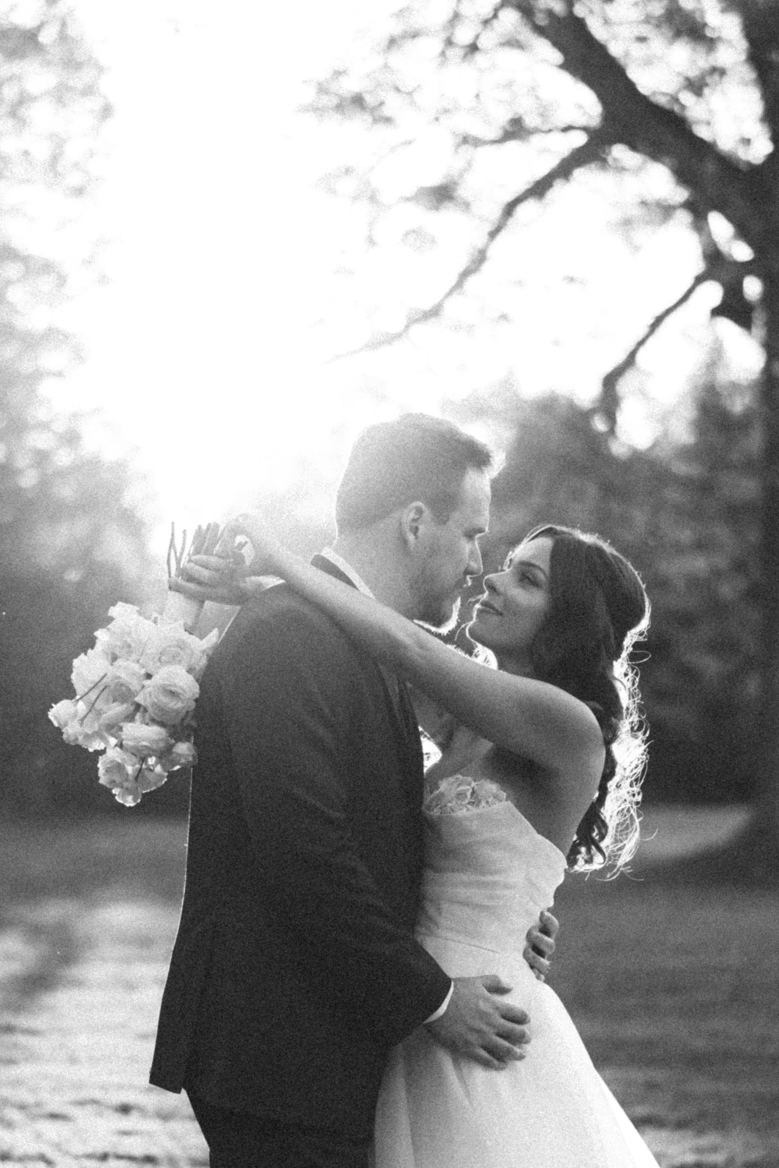 A black and white photo of a bride and groom embracing outdoors, with the bride holding a bouquet of flowers and sun shining behind them.