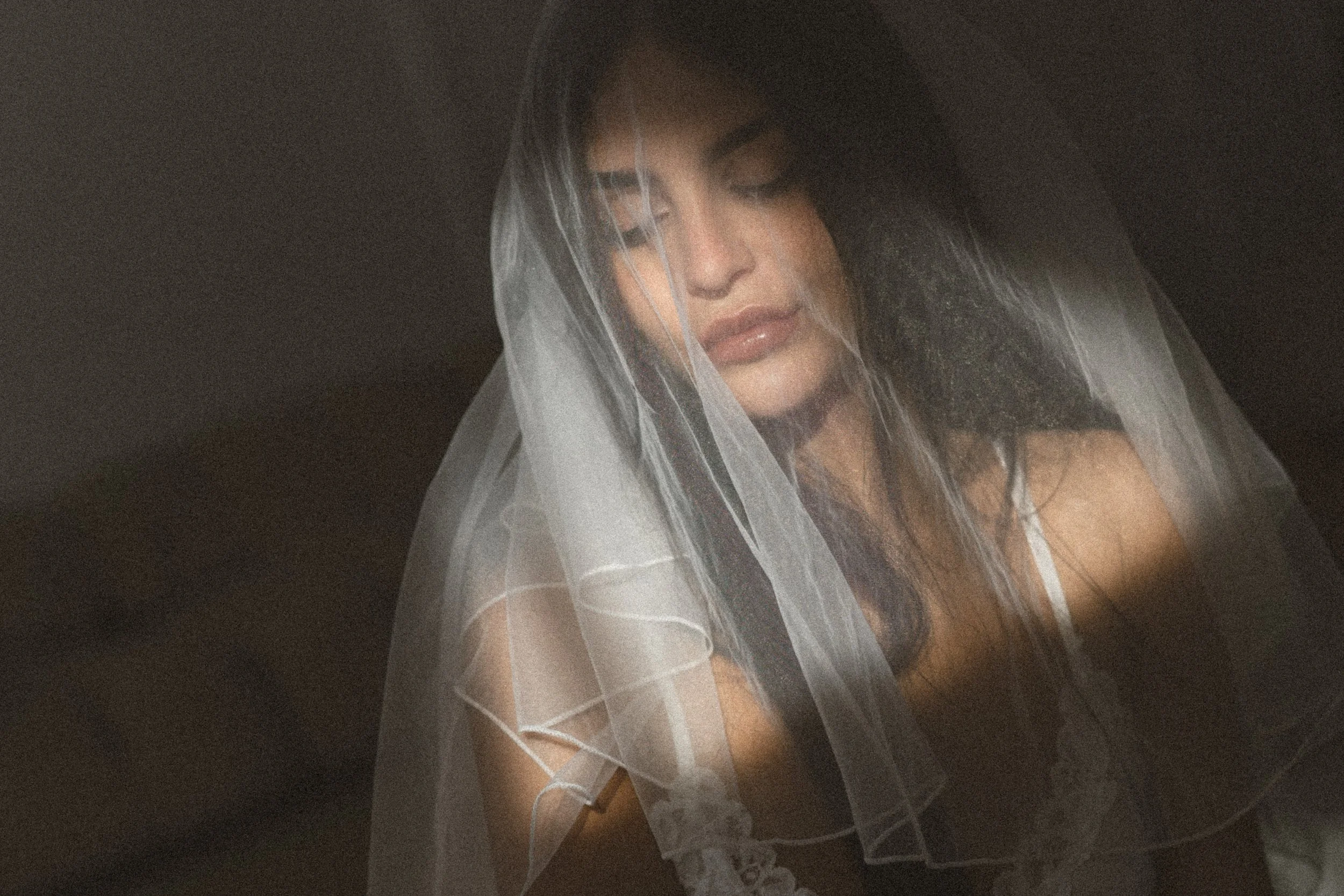 A woman with dark hair and light makeup, wearing a sheer white veil and lace lingerie, looks down with a contemplative expression, against a dark background.