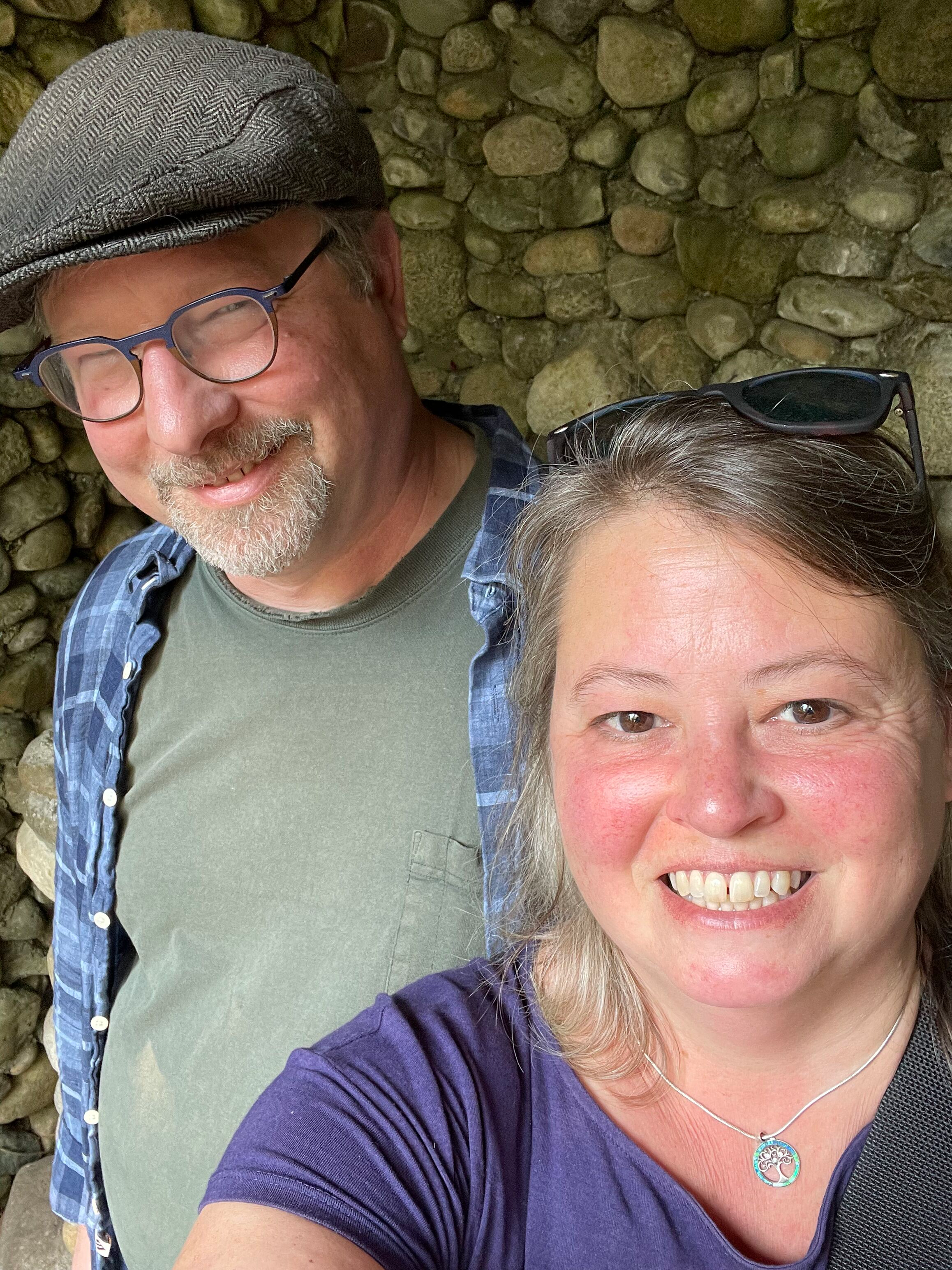 A smiling man wearing glasses, a gray cap, a blue plaid shirt, and a green t-shirt, and a woman with sunglasses on her head, smiling, wearing a purple shirt and a necklace with a tree pendant. They are standing in front of a rocky stone wall.