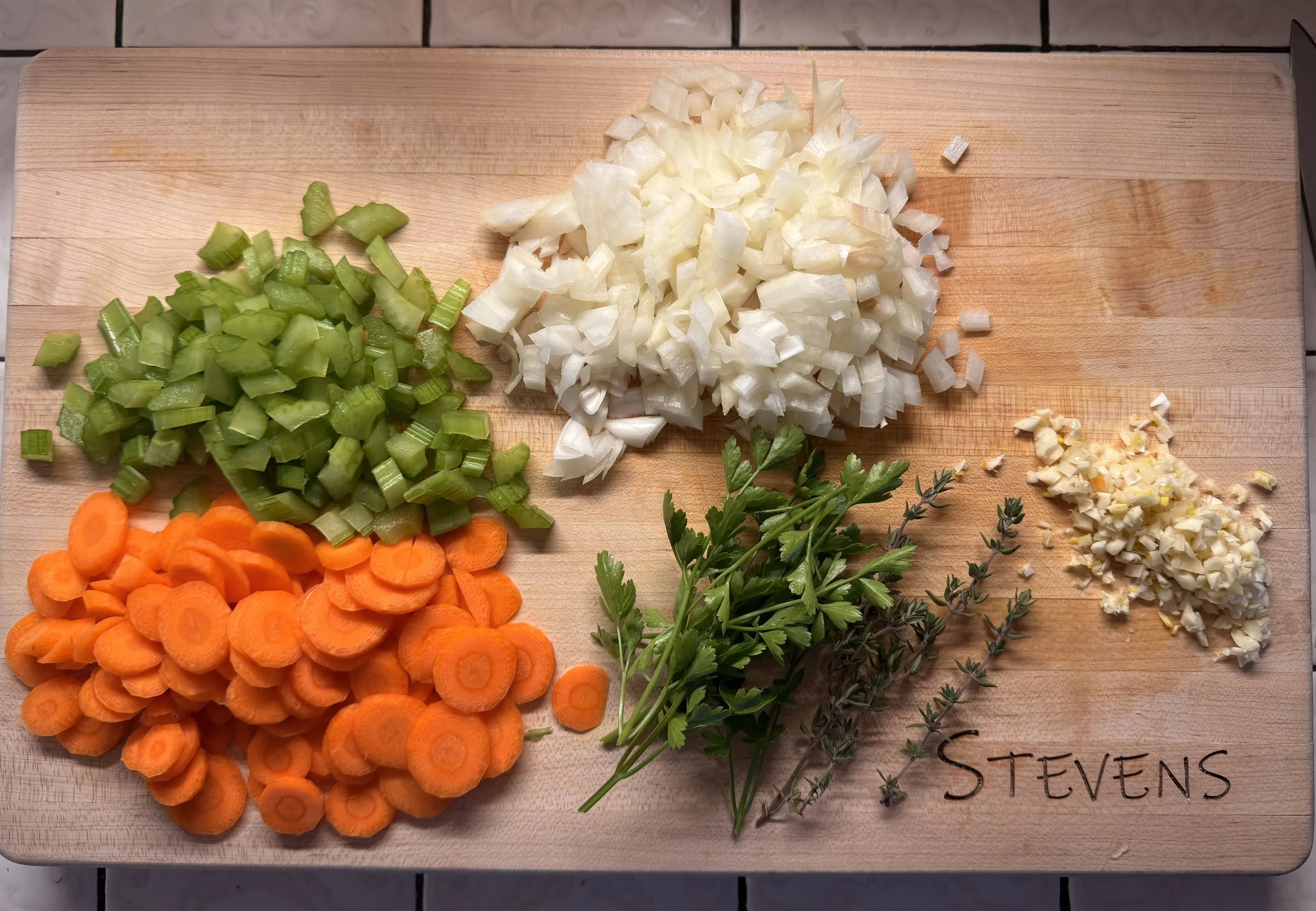 Chopped celery, onion, garlic, parsley, thyme, and sliced carrots on a wooden cutting board.