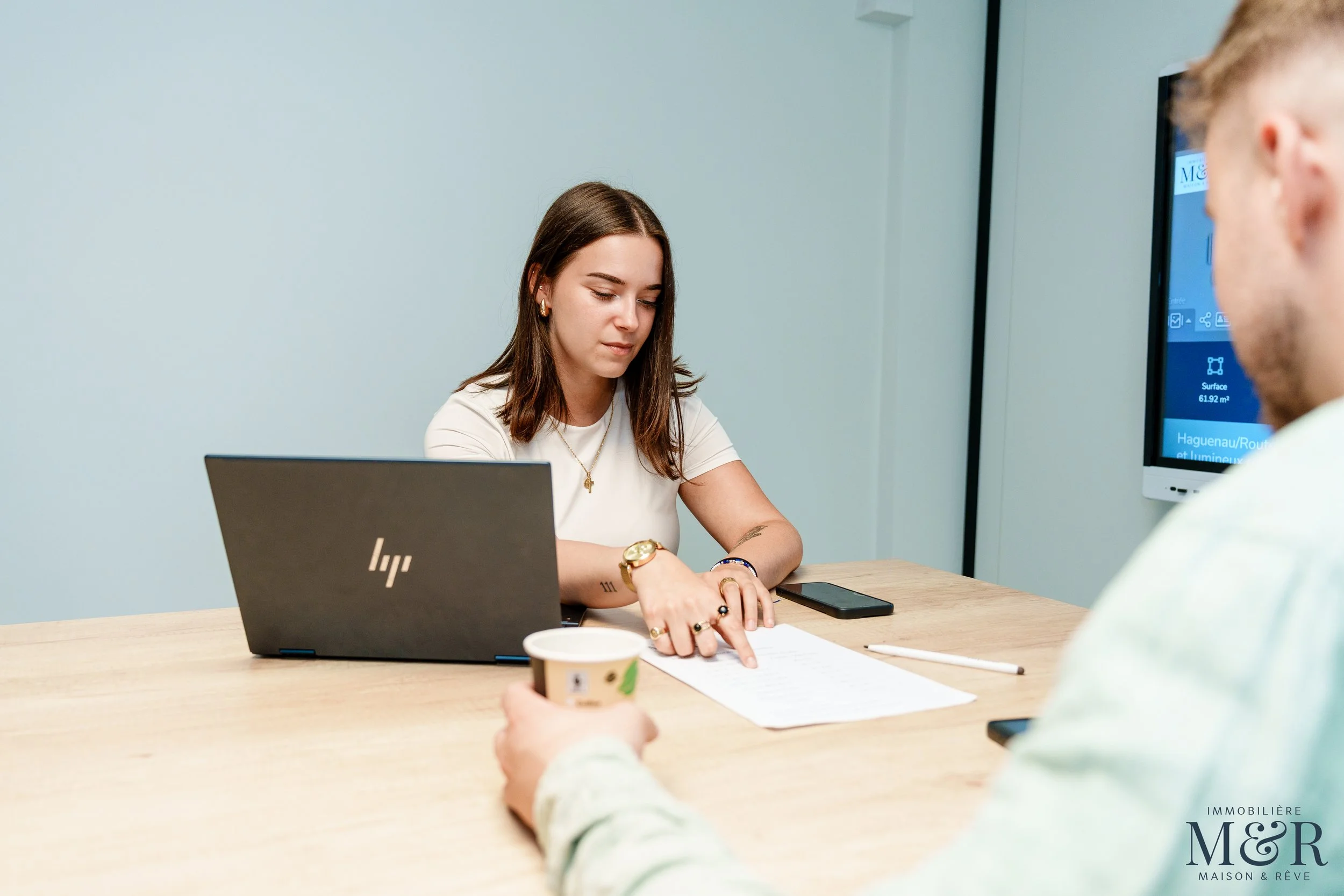Une femme et un homme lors d'une réunion de travail en salle de conférence, la femme indique un document avec un stylo, l'homme tient une tasse de café.