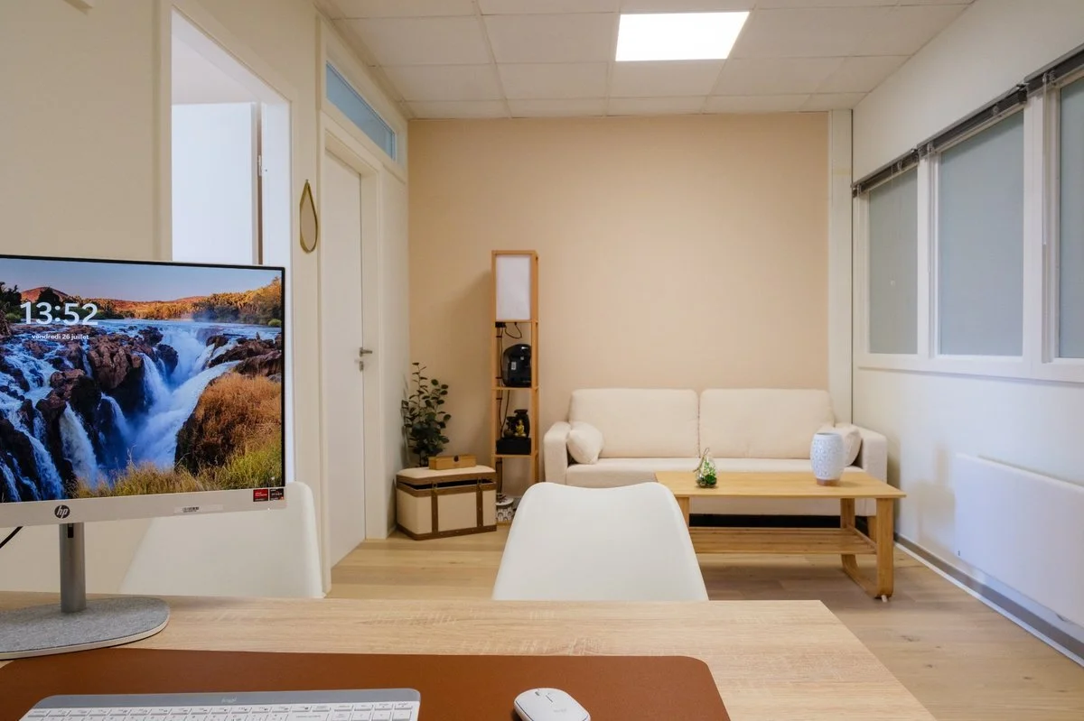 Salon avec canapé beige, table en bois, étagère en bois avec objets décoratifs et plantes, grandes fenêtres avec stores, écran d'ordinateur sur table en bois, bureau avec clavier et souris, plafond à carreaux avec lampes LED.