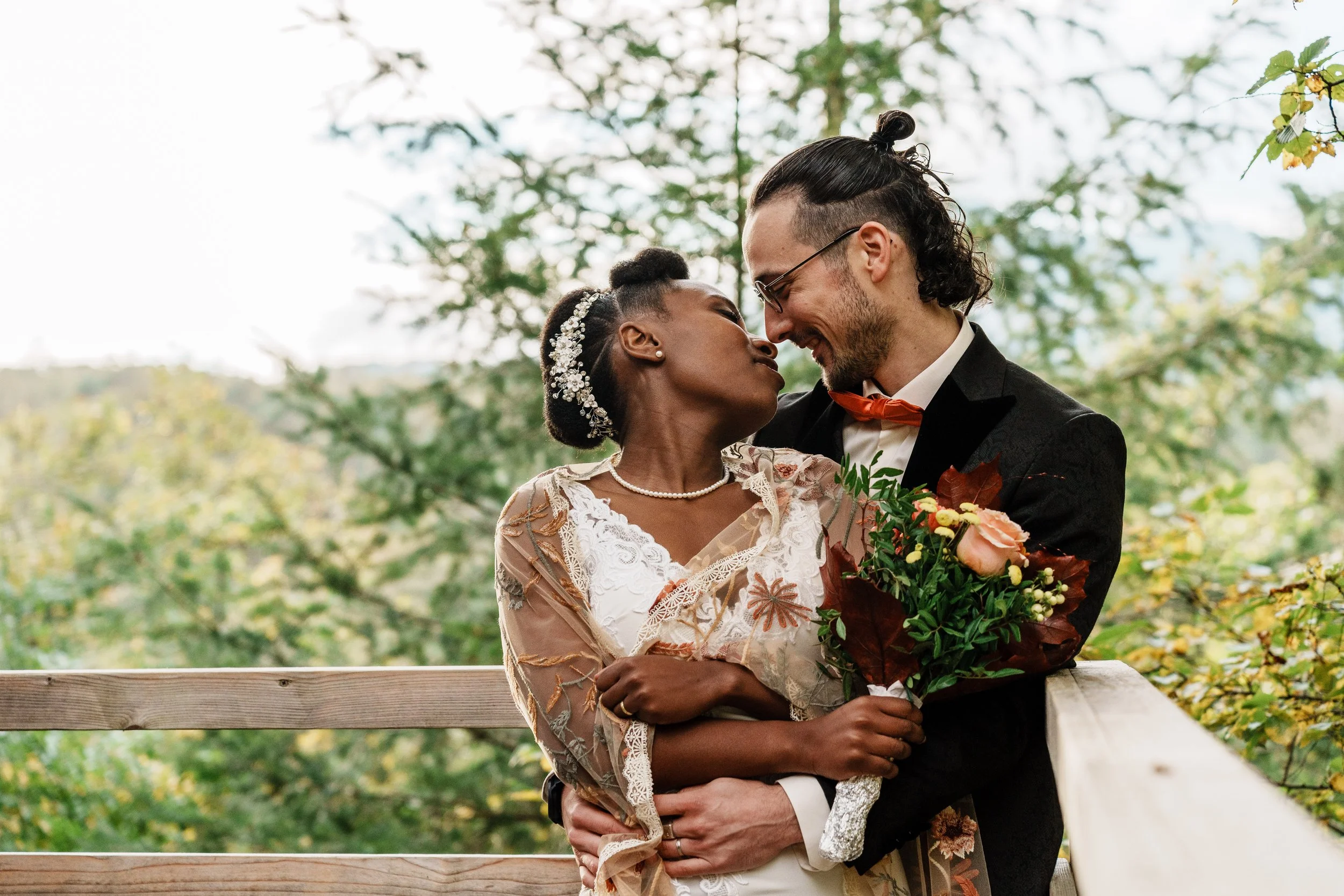 Un couple de mariage s'embrassant, tenant un bouquet de fleurs, dans un cadre extérieur avec des arbres en arrière-plan.