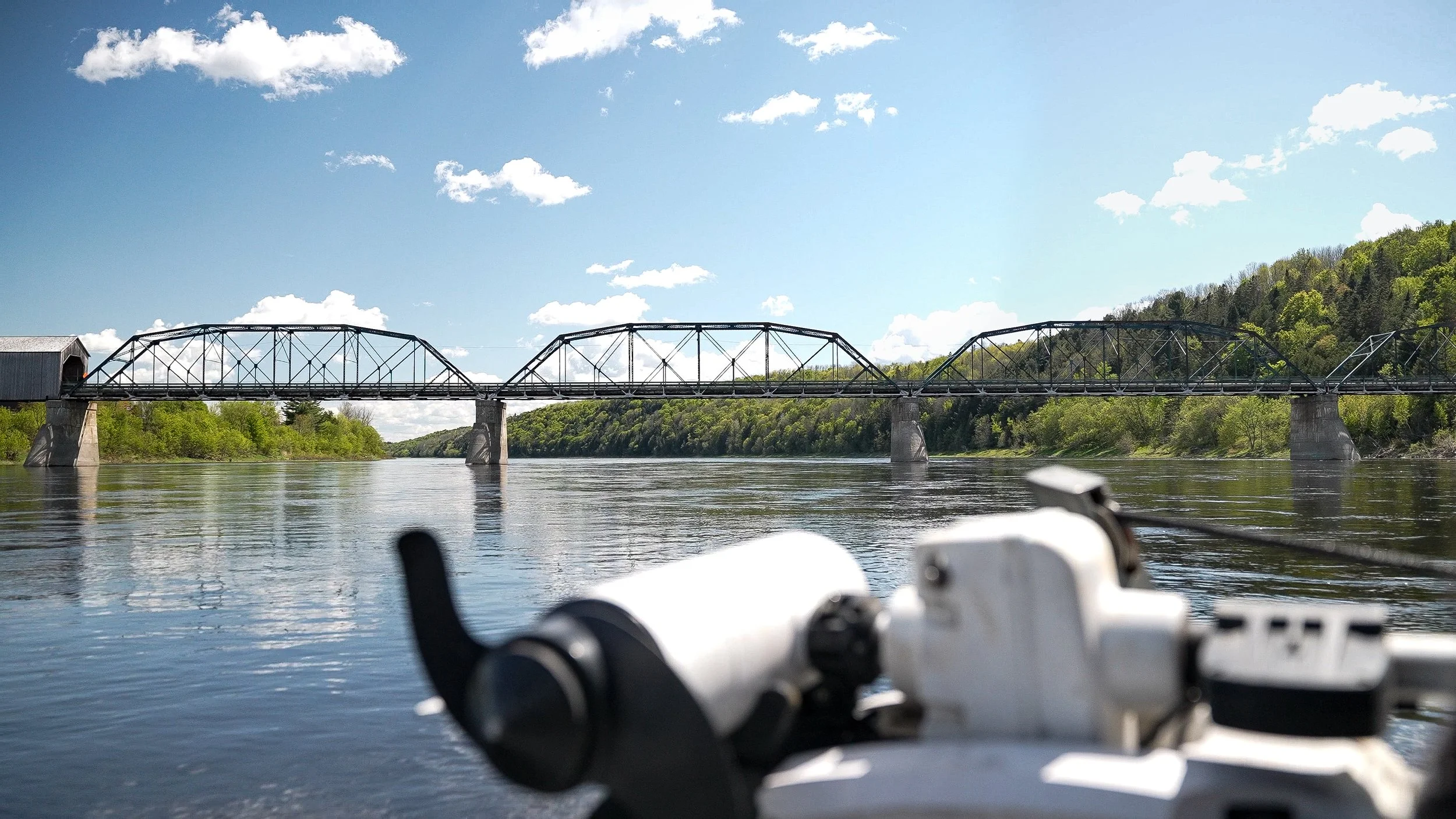 View of a bridge over a river with a bicycle handlebar in the foreground, lush green trees on both sides, blue sky with scattered clouds.