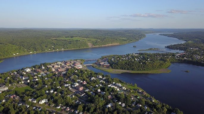 Aerial view of a riverside town with houses, roads, and a bridge crossing a wide river, surrounded by green hills and water bodies.