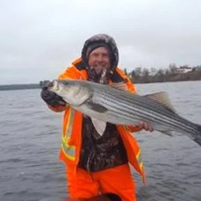 A person dressed in waterproof gear holding a large fish outdoors near a lake