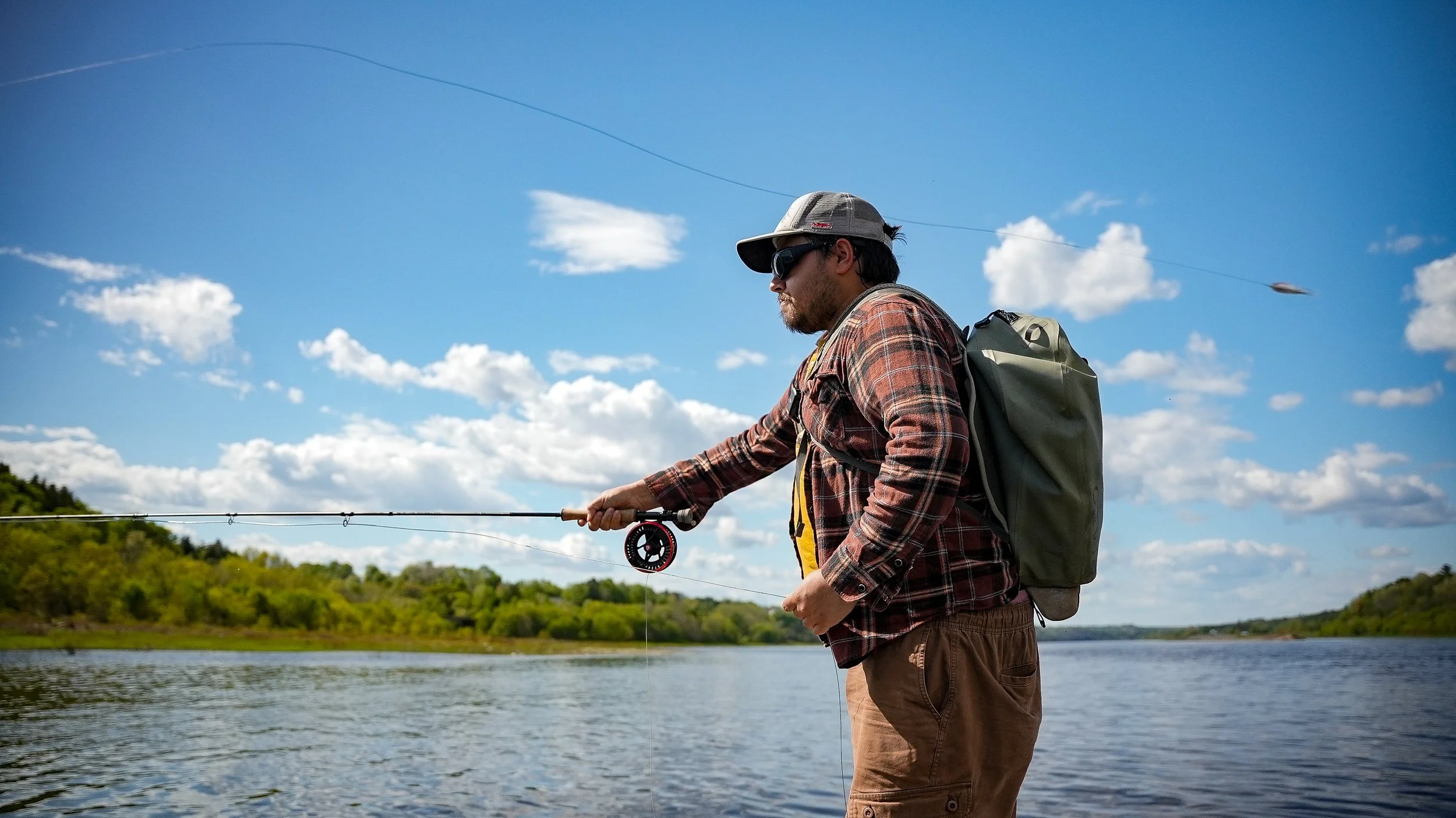 Man fishing in a river on a bright, partly cloudy day wearing sunglasses, a cap, a plaid shirt, and a backpack.