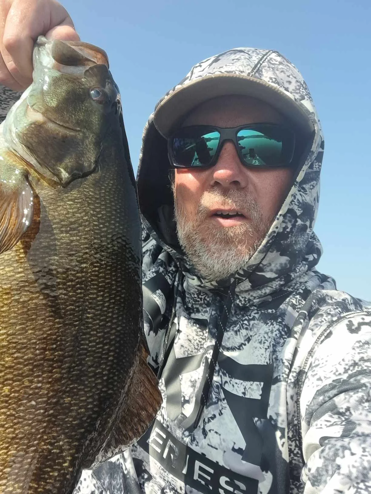 Man in patterned hoodie and sunglasses holding a large fish outdoors against a clear blue sky.