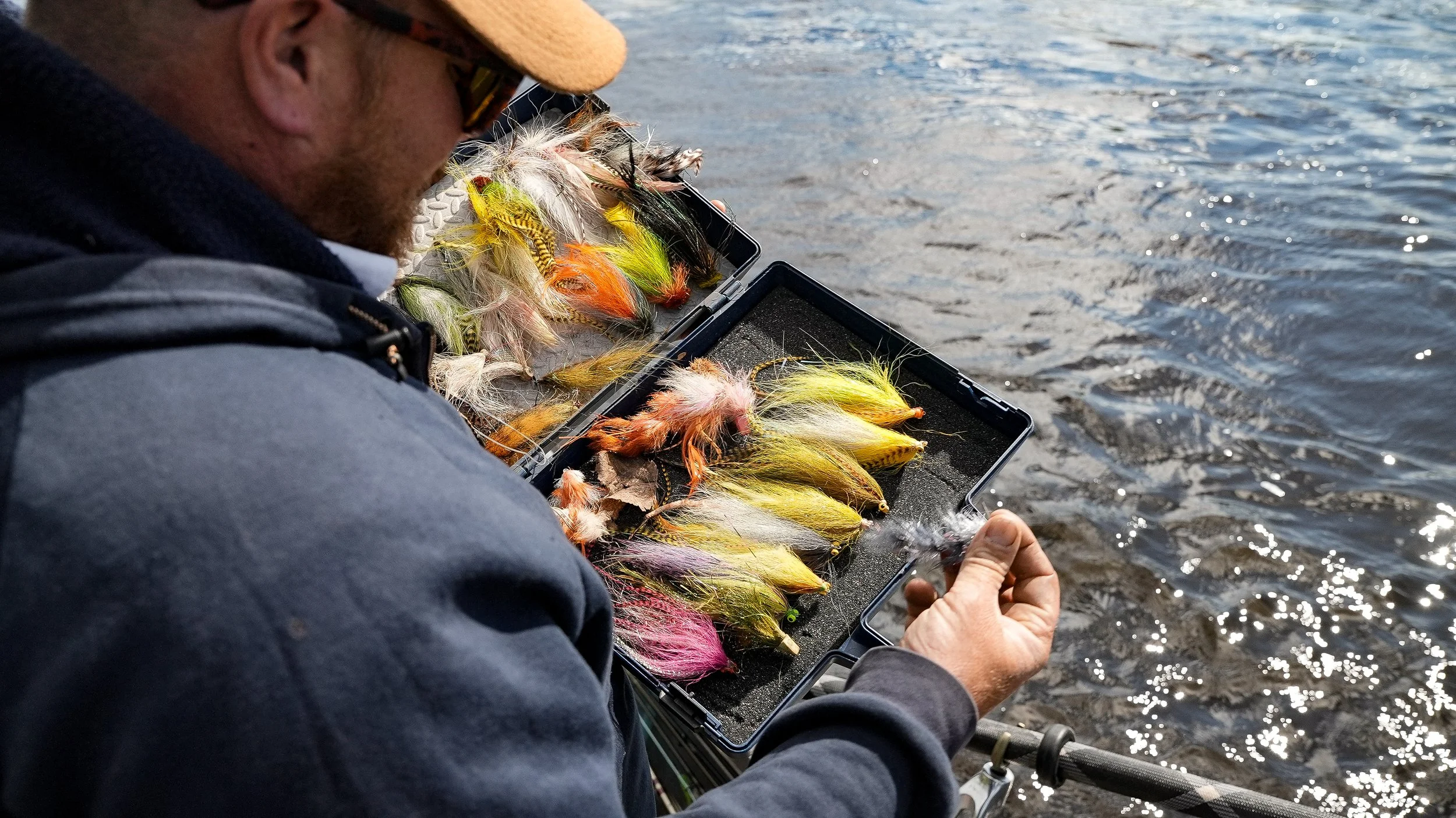 A man in a hoodie and hat arranging colorful fishing flies in a tackle box by the water on a fishing rod.