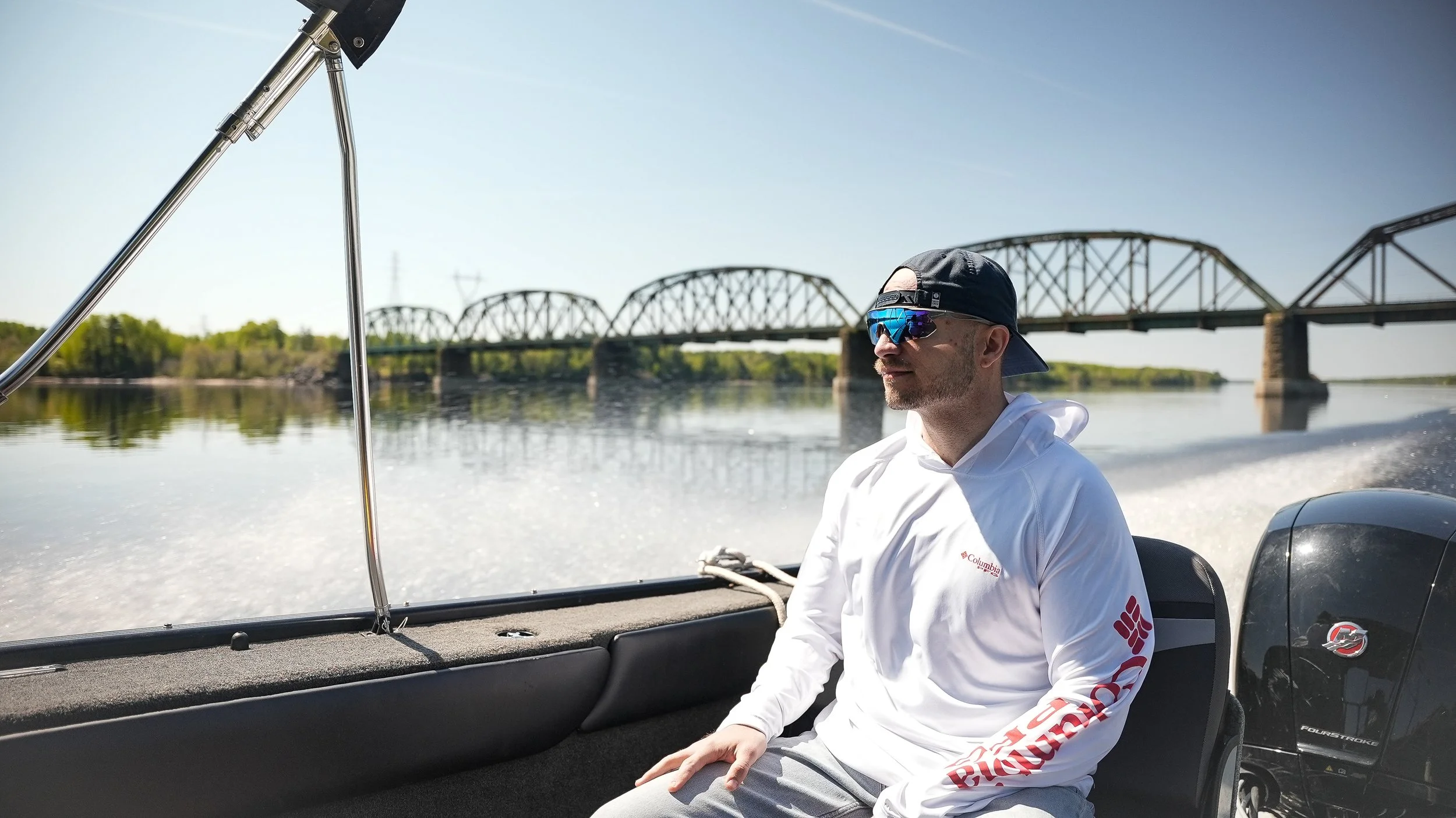 A man wearing sunglasses, a baseball cap, and a white Columbia hoodie is sitting in a boat on a river, with a bridge visible in the background.