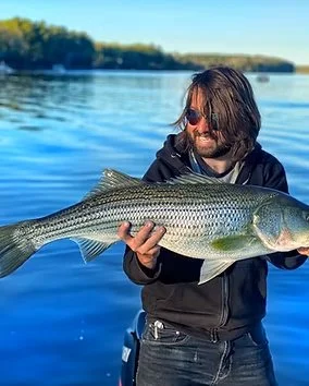 Person with shoulder-length hair and sunglasses holding a large fish near a body of water.