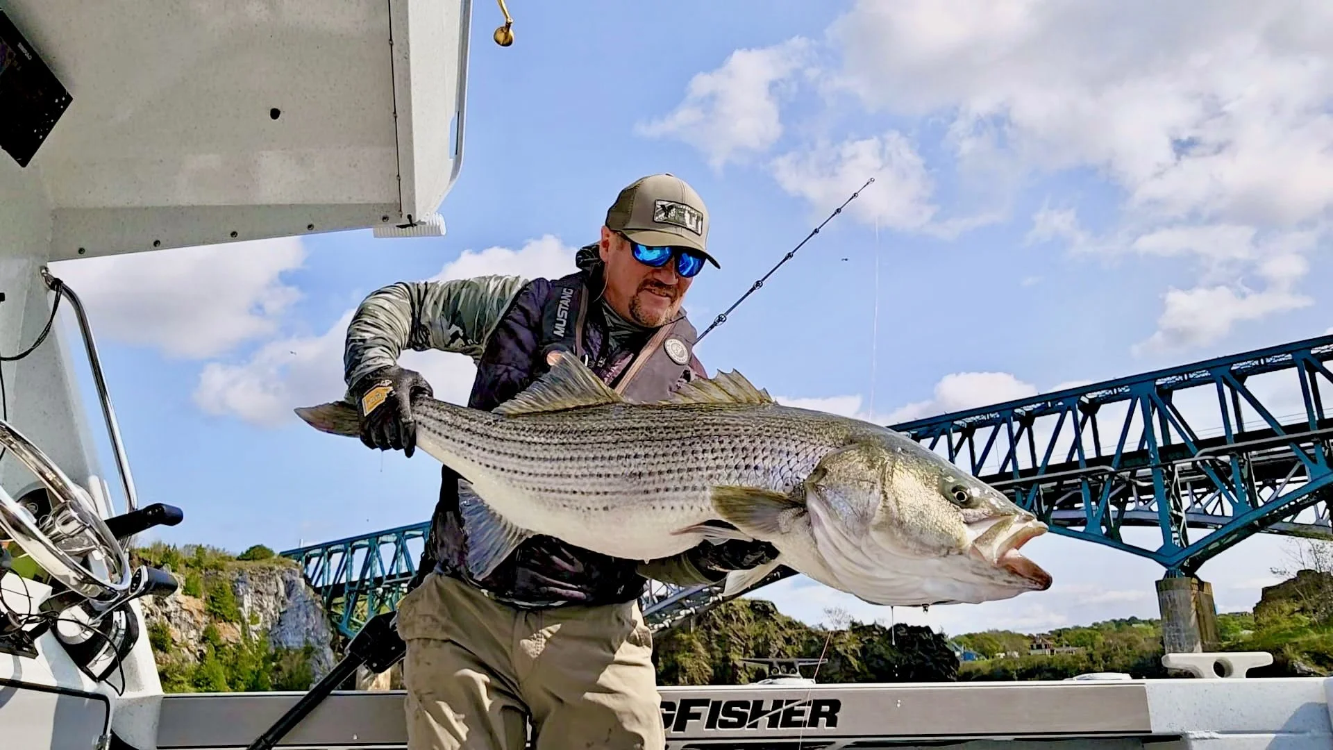 Man wearing sunglasses and a cap holding a large striped bass on a boat, with a bridge and blue sky with clouds in the background.