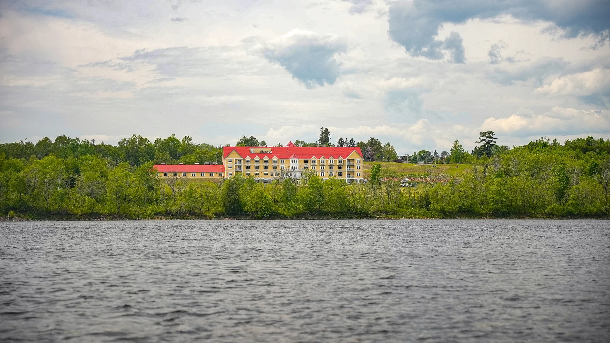 A large yellow building with a red roof on a hill, surrounded by green trees, next to a body of water with a cloudy sky above.