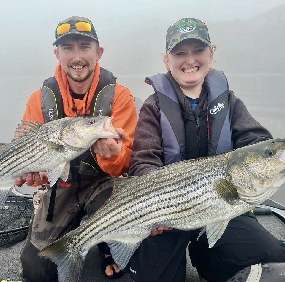 Two people holding large striped bass fish on a boat, with foggy water in the background.