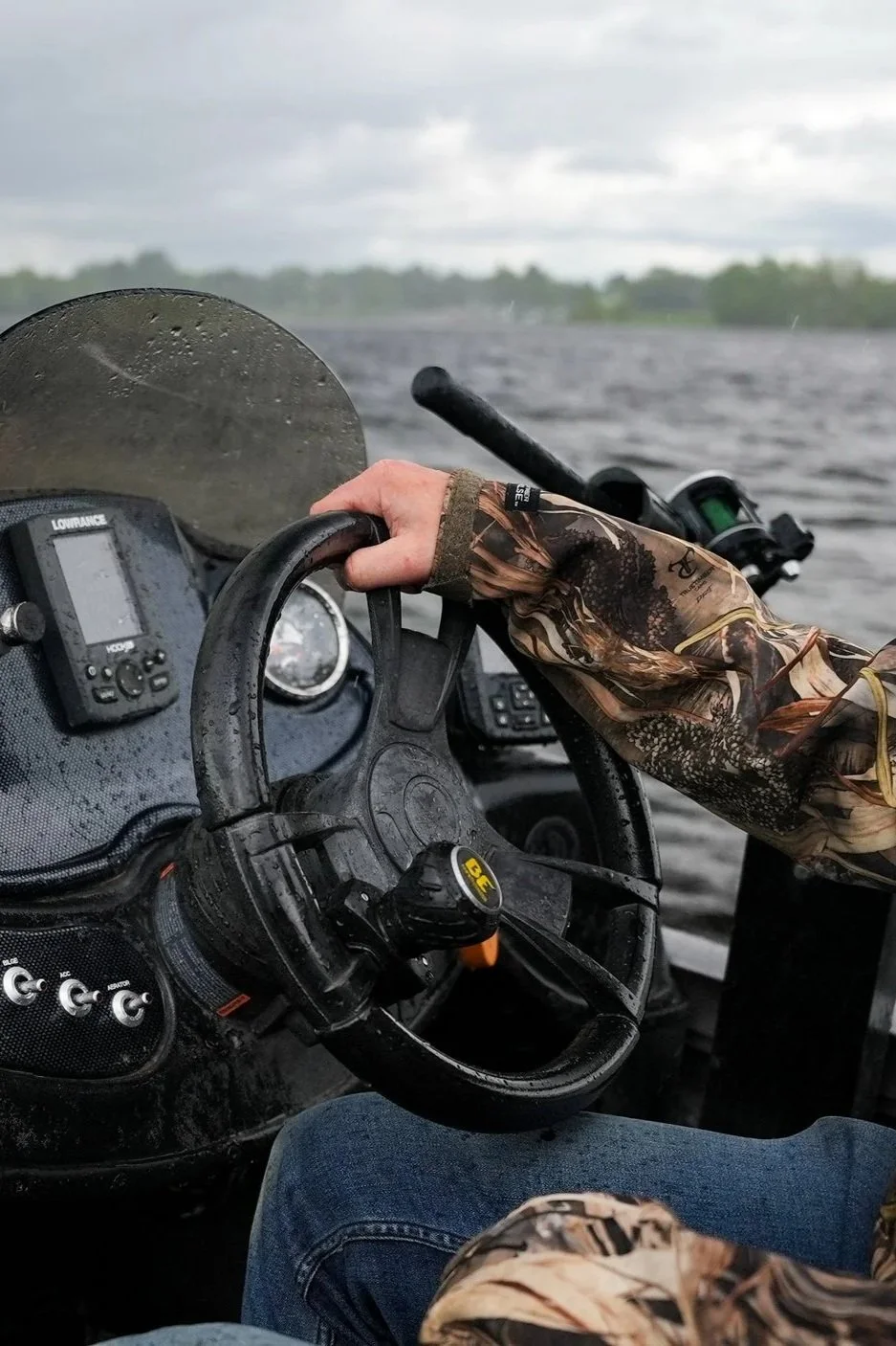 A person driving a boat in choppy water on a cloudy day, wearing camouflage clothing and holding the steering wheel.