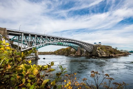 A bridge over a river under a partly cloudy sky, with greenery in the foreground.