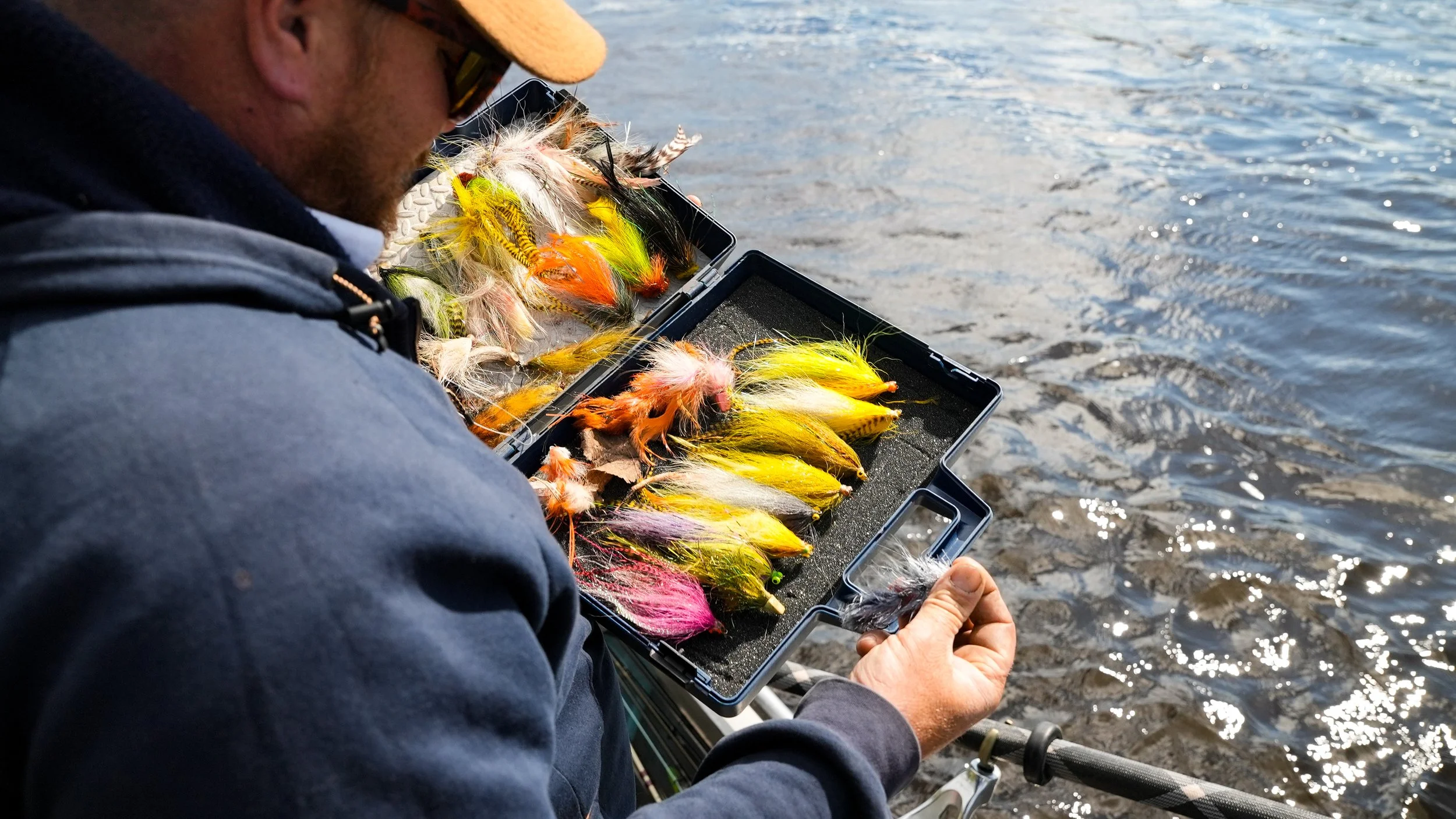 A man holding a box of fishing lures with colorful feathers while sitting by the water.