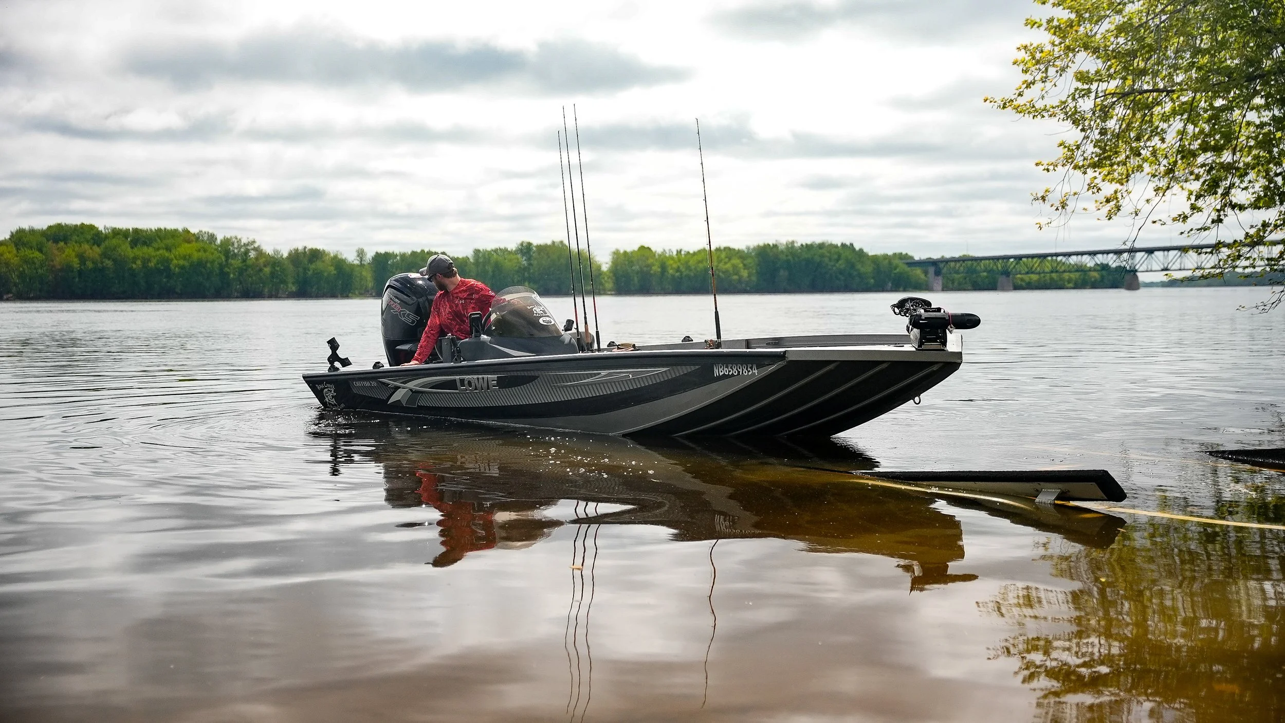 A person in a red jacket standing on a small black fishing boat on a lake, holding fishing rods, with trees and a bridge in the background under cloudy skies.