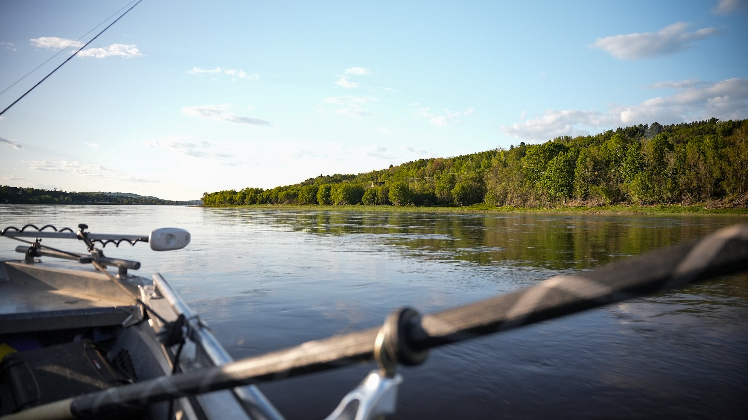 View of a river landscape from a boat, with the bow and fishing equipment visible in the foreground, and green trees along the riverbank under a partly cloudy sky.