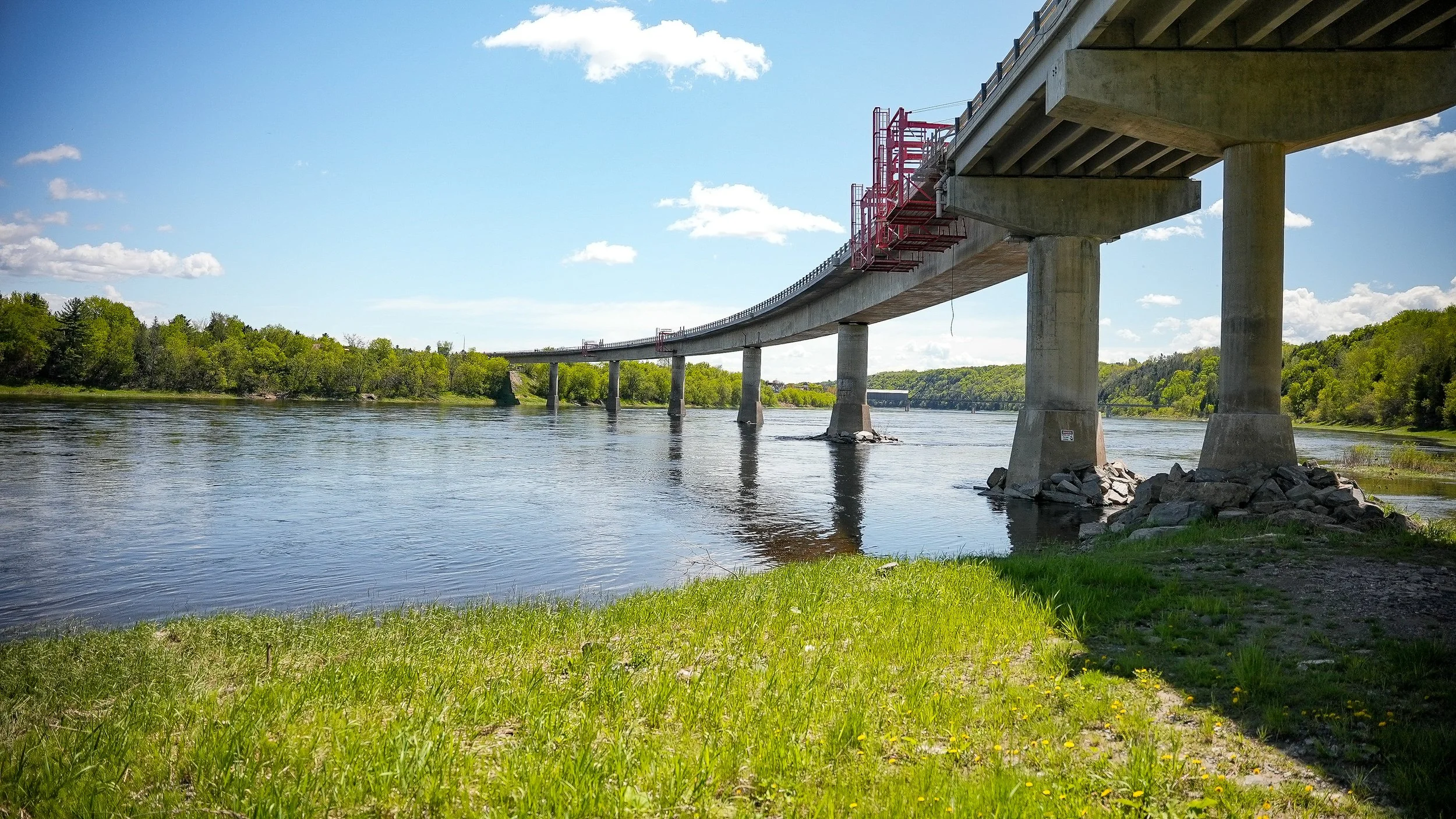 Bridge over a river with green trees on the riverbank and a grassy area in the foreground on a sunny day with a blue sky and clouds.
