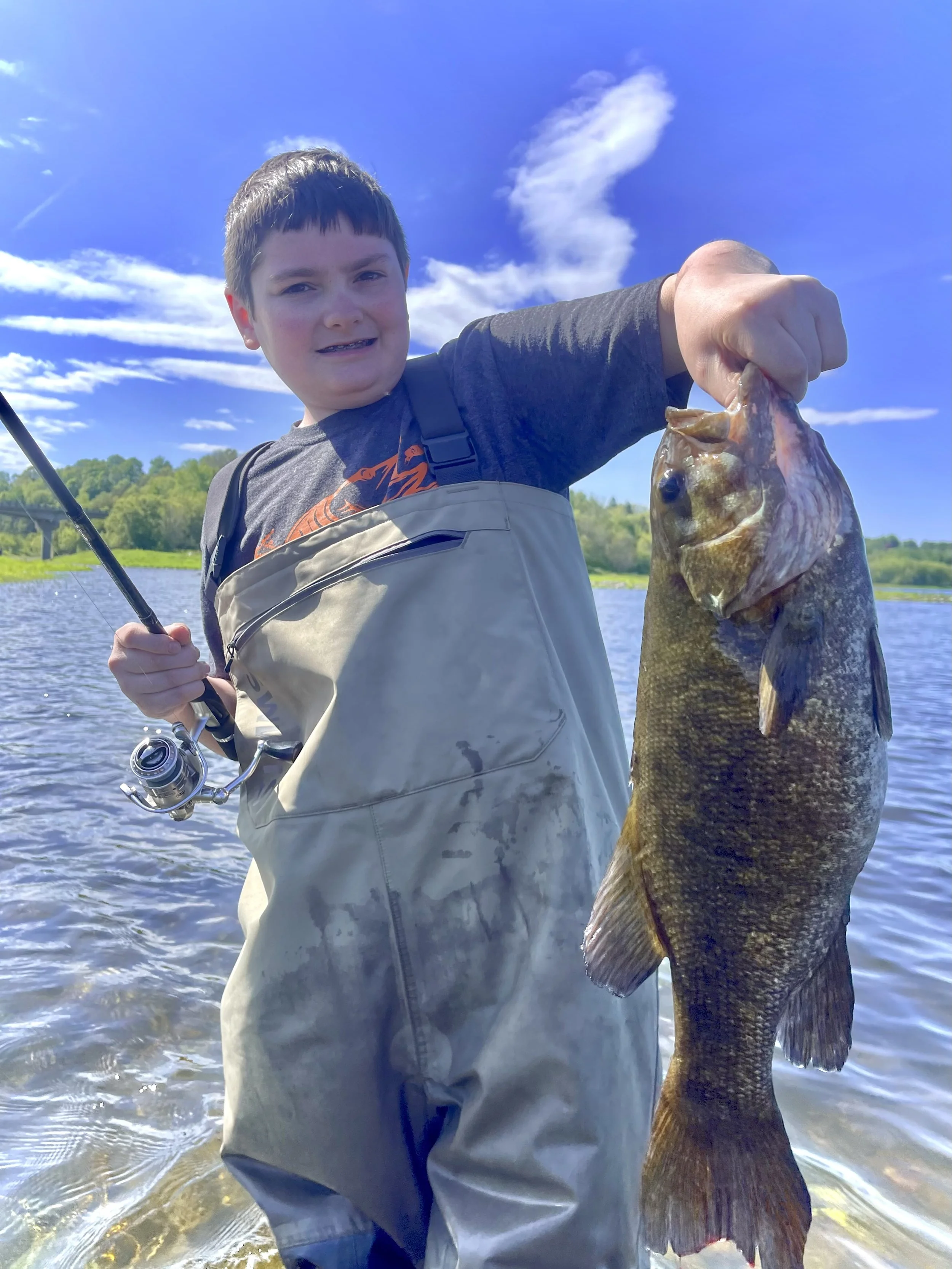 A man holding a large fish on a boat in a river with forested hills in the background.