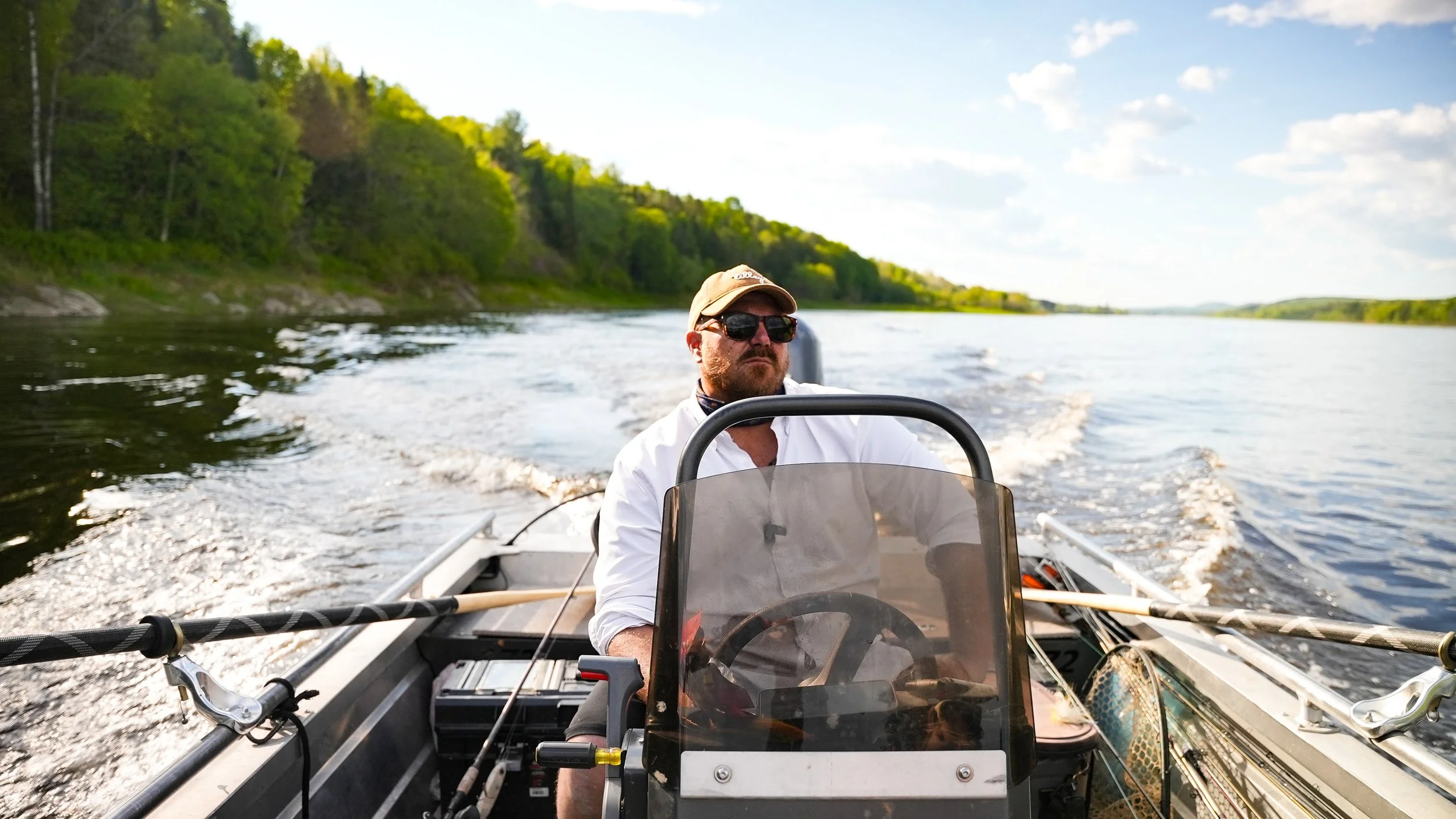 Man wearing sunglasses and a cap steering a boat on a river with green trees and a blue sky in the background.