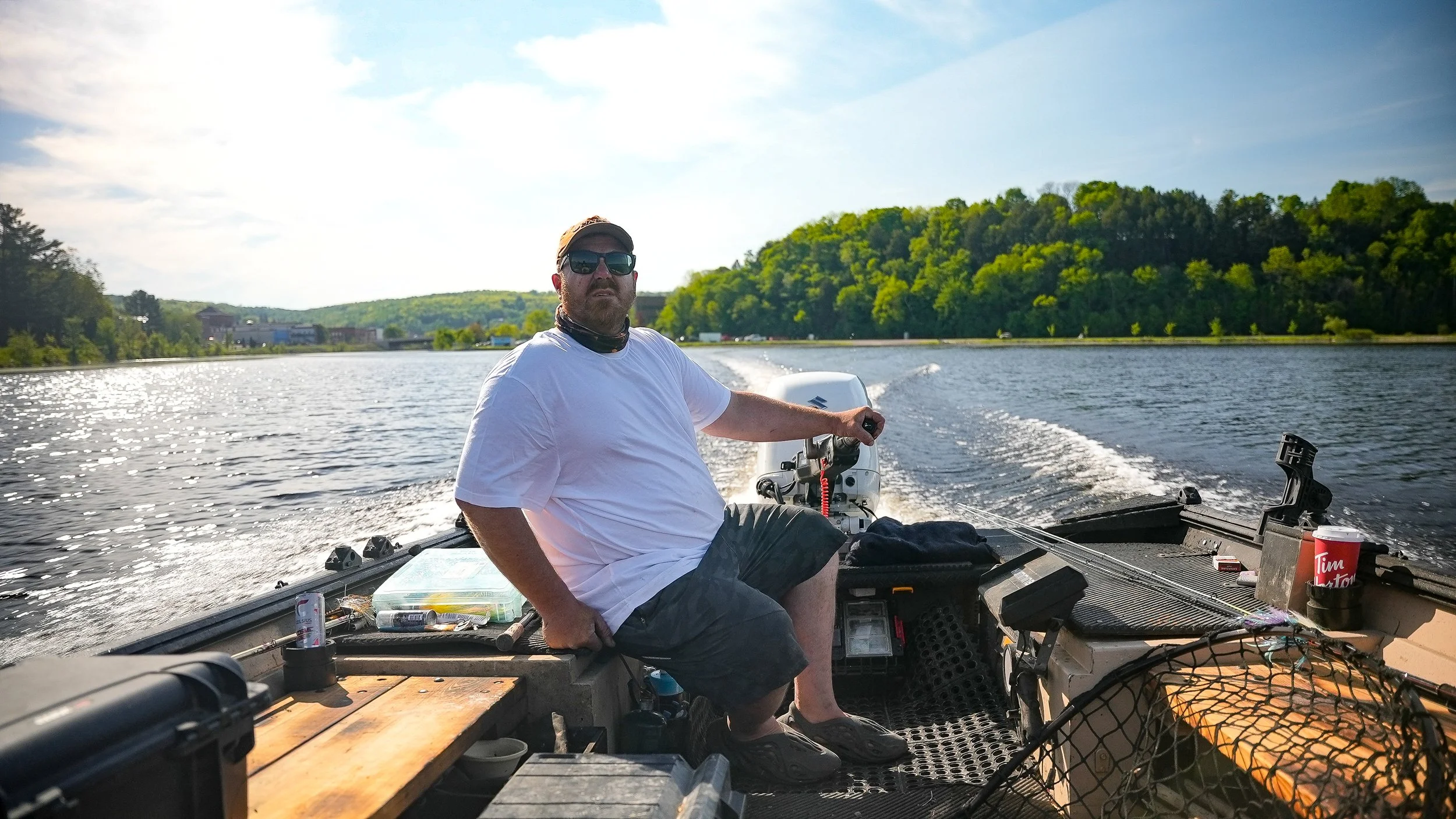 Man sitting on boat with outboard motor on a river, surrounded by green trees, wearing sunglasses, white t-shirt, and shorts, with water bottles and fishing gear nearby.