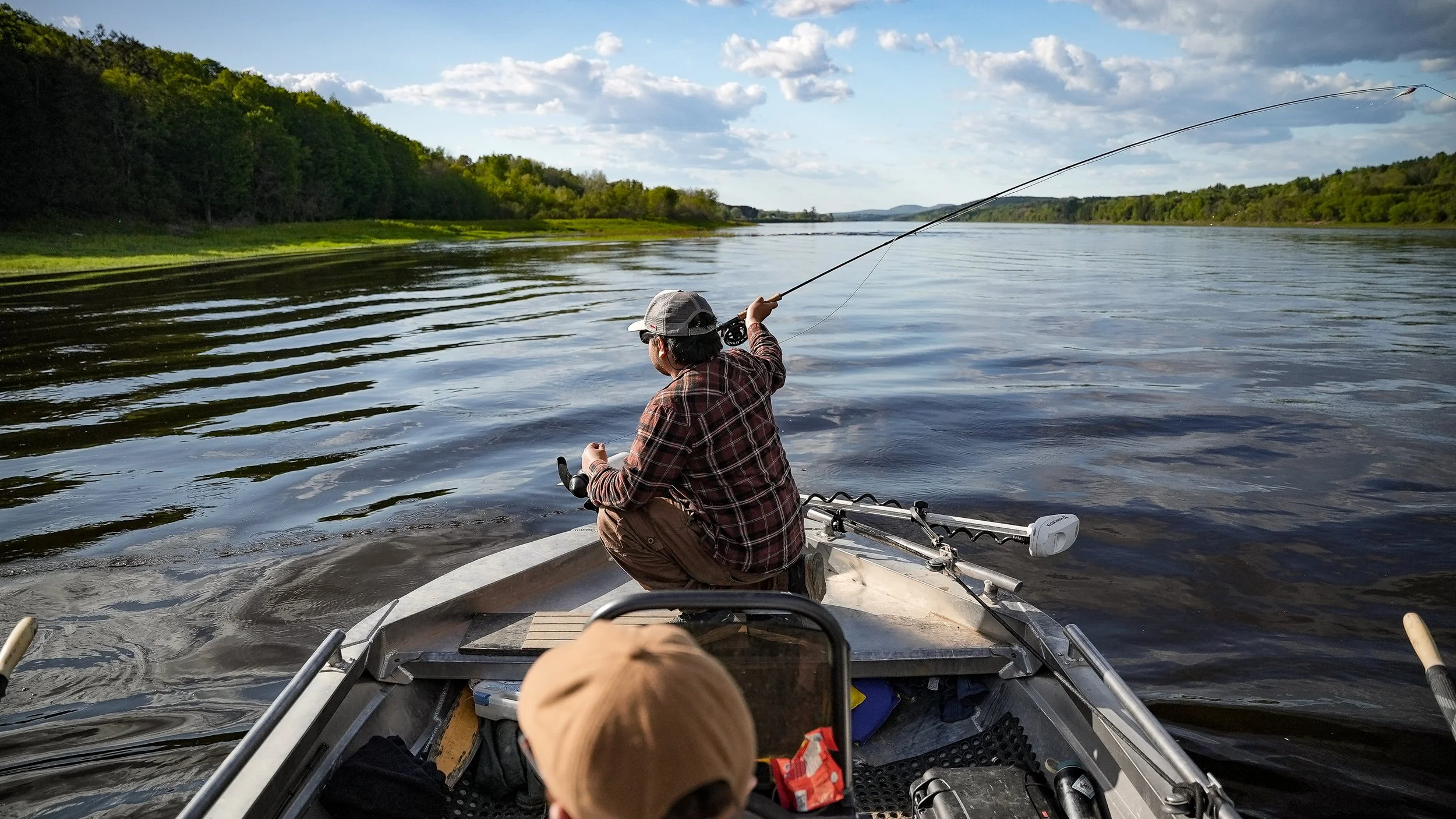Person fishing from a boat on a calm river with green trees on the riverbank and a partly cloudy sky