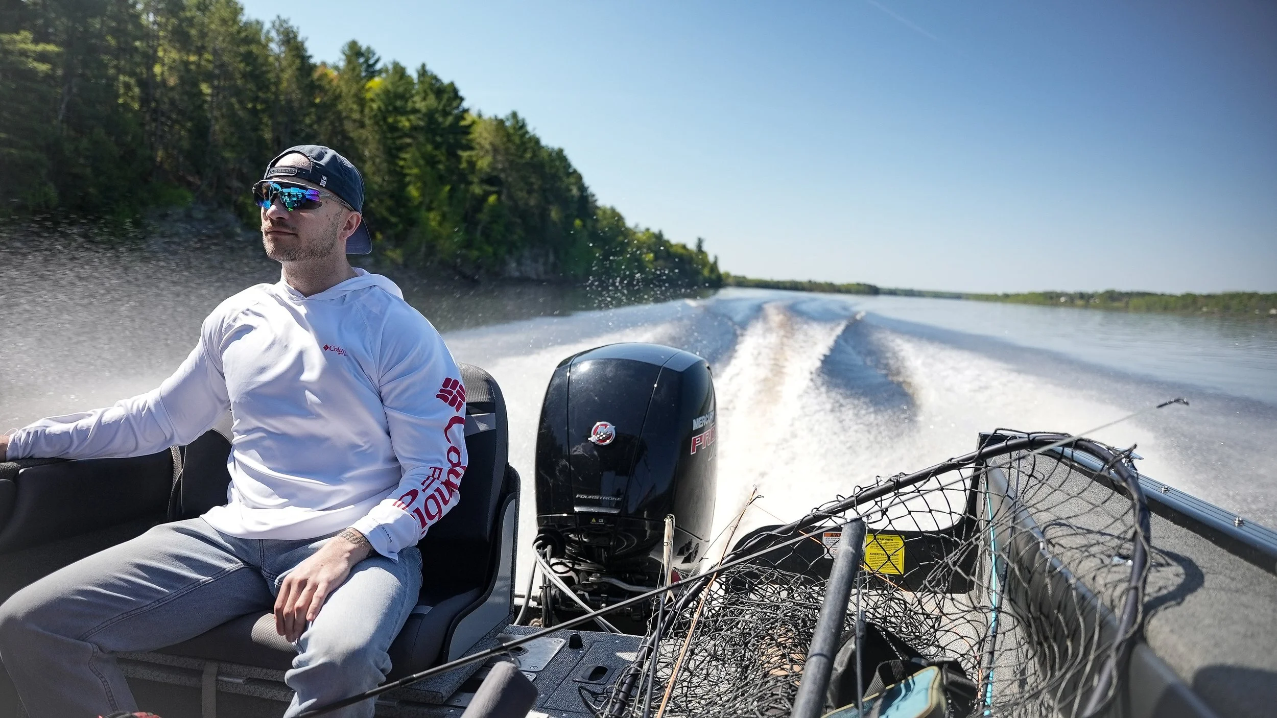 Man wearing sunglasses and a white hoodie is sitting on a boat with a black outboard motor, cruising on a wide river with trees on the shoreline under a clear blue sky.
