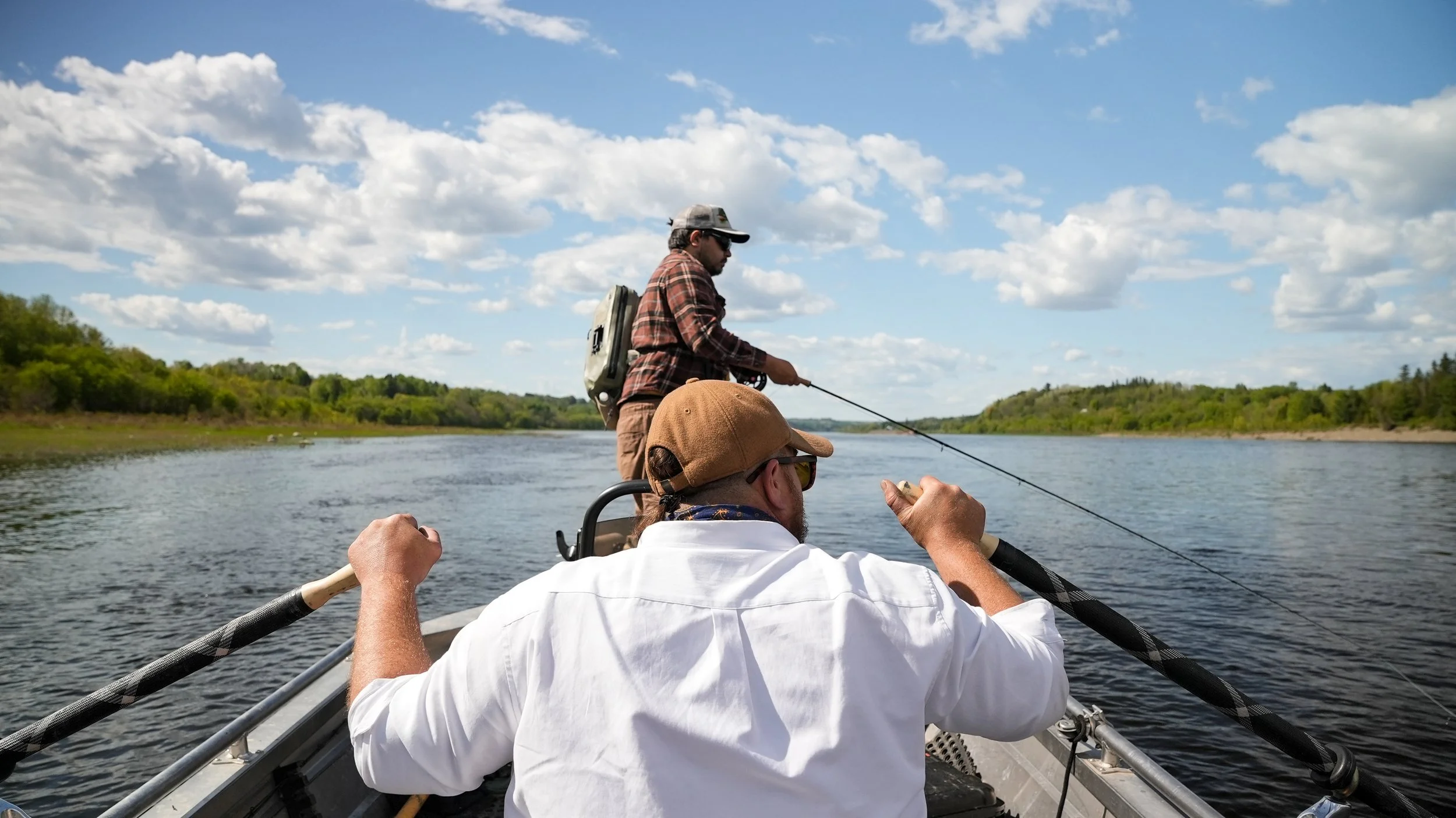 Two men fishing from a boat on a wide, calm river. One man is standing, casting his fishing line, while the other is sitting at the back of the boat, holding an oar. The scene is set on a sunny day with a partly cloudy sky and green trees on the rive