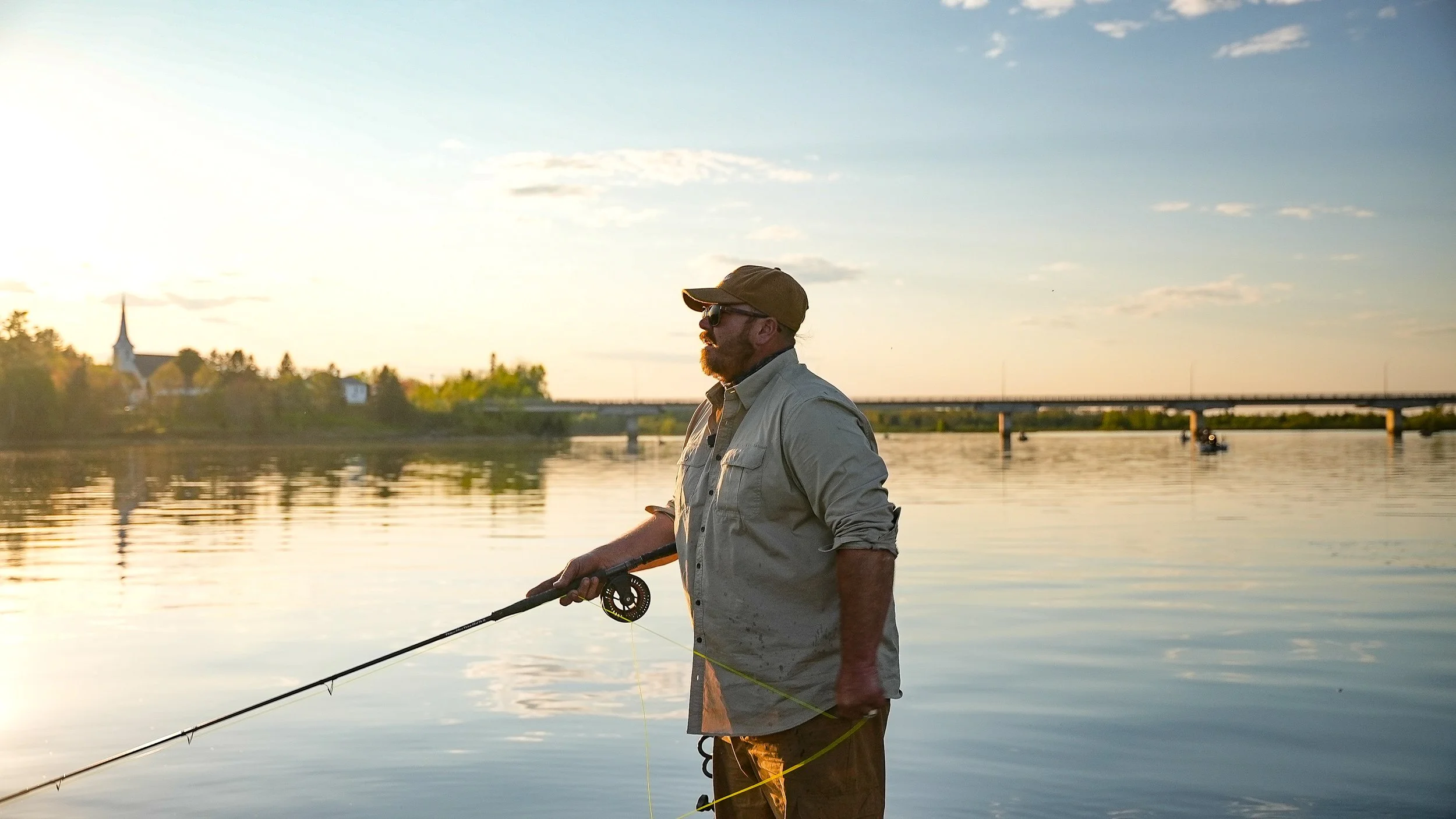 Man fishing in a boat on a calm river during sunset with a bridge and trees in the background.