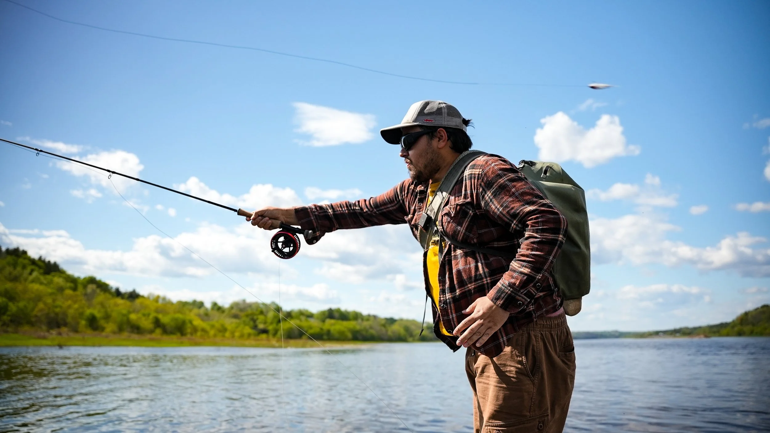 Man fishing on a river, wearing a plaid shirt, cap, sunglasses, and carrying a backpack.