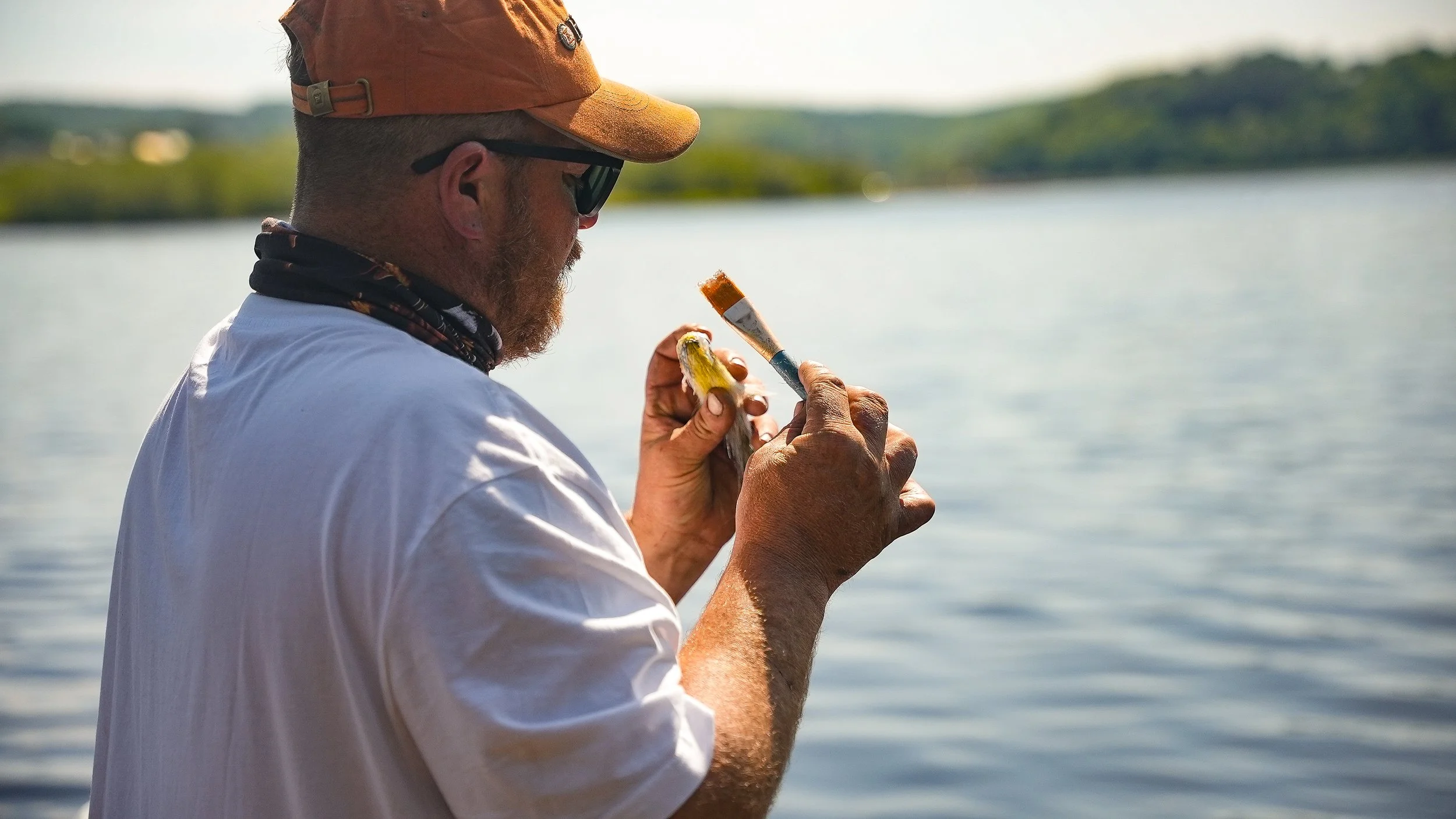 A man with a beard, wearing a brown cap, sunglasses, a white shirt, and a neck gaiter, painting a small object near a body of water with a brush.