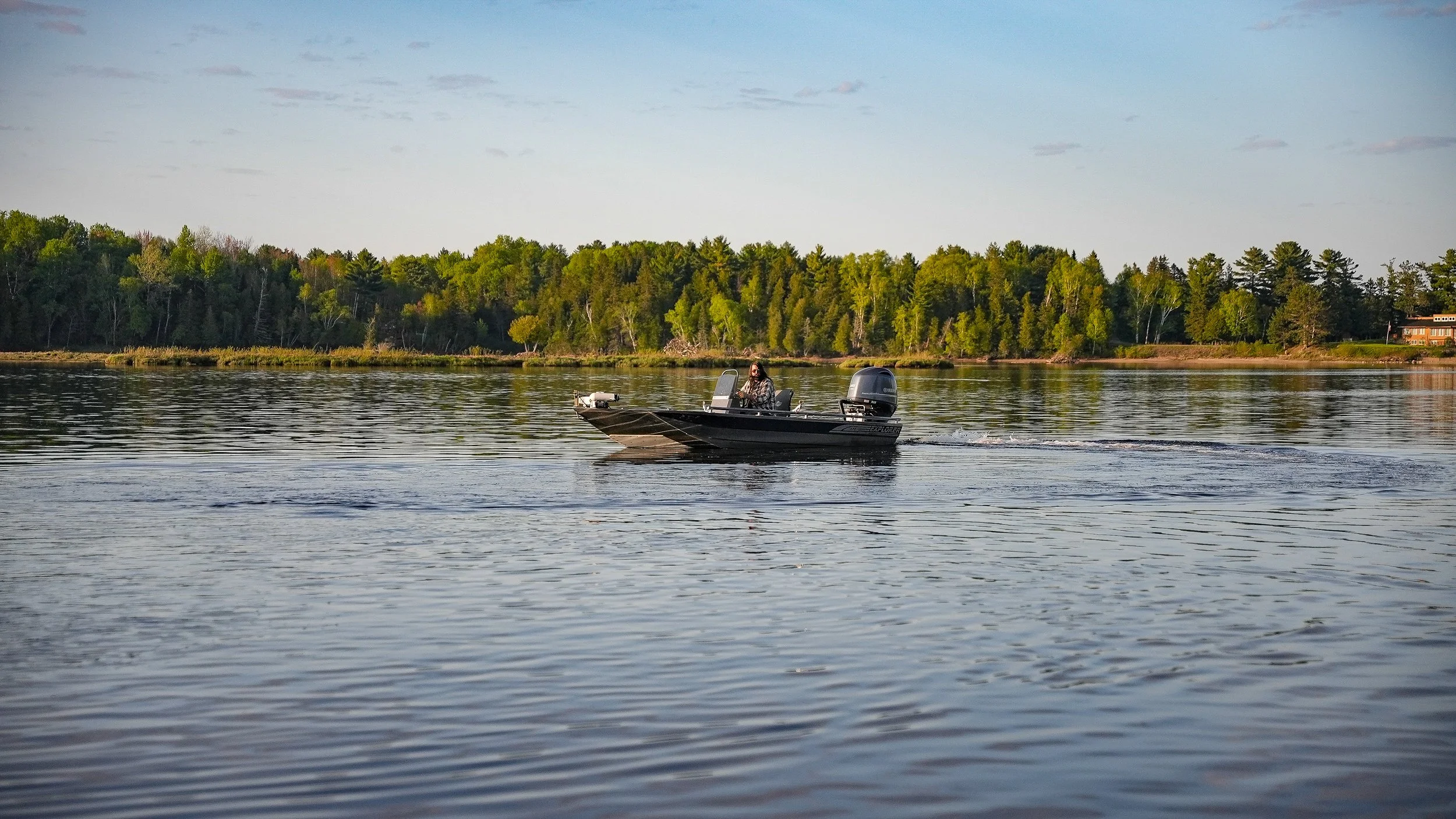 A person steering a small motorboat on a calm body of water with a tree-lined shoreline in the background during daylight.