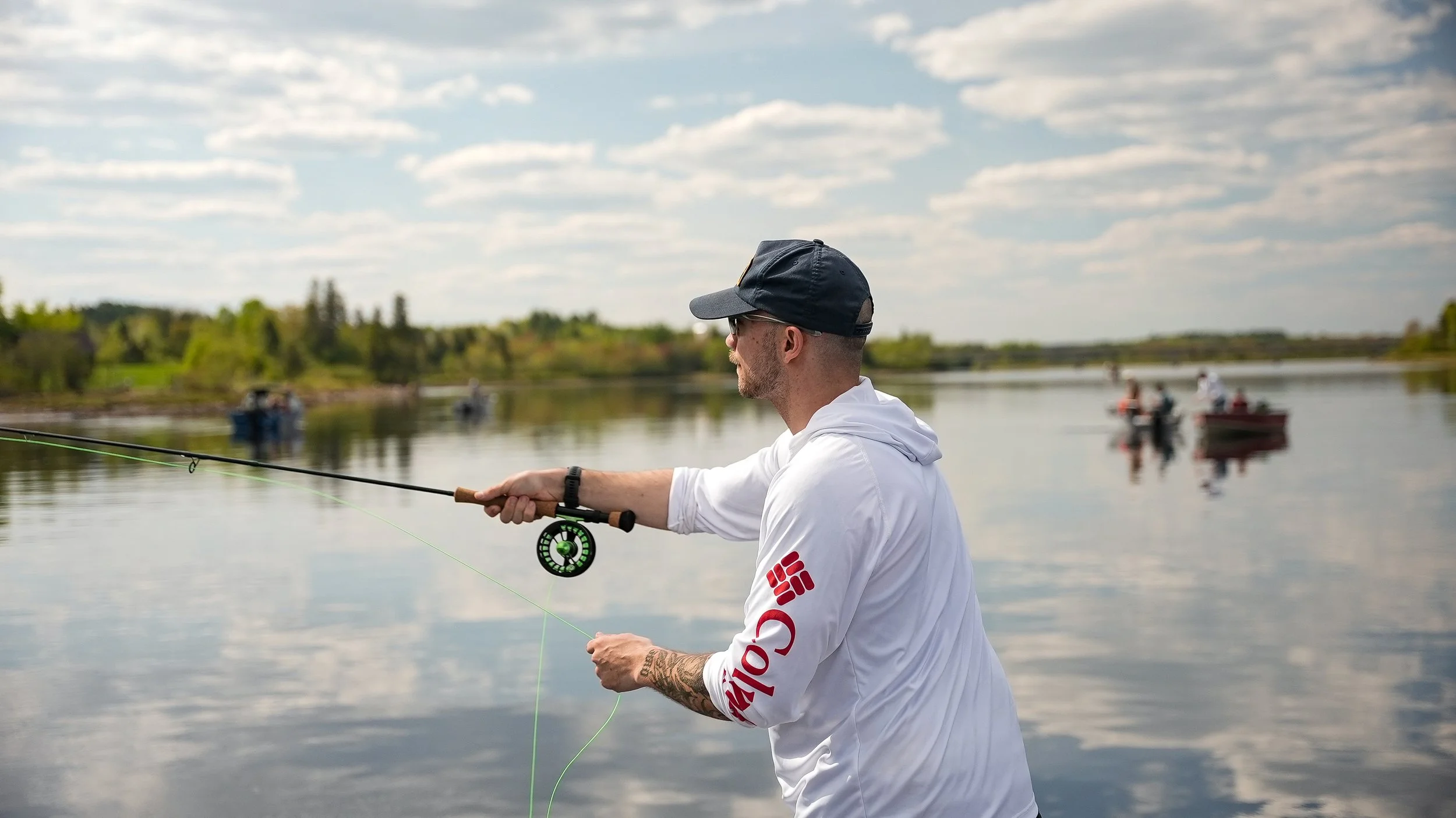 A man fishing by a calm lake on a partly cloudy day, with several boats visible in the background.